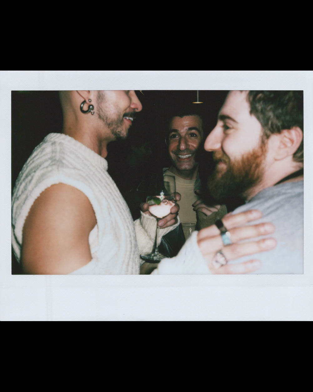 Three men smiling and socializing indoors, one holding a glass with a drink, in a dimly lit setting.