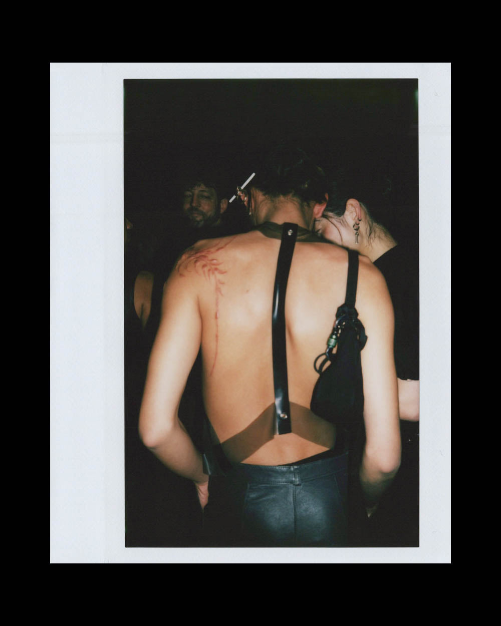 Person with a bare back wearing leather harness and pants, standing next to another person in dark clothing in low light.