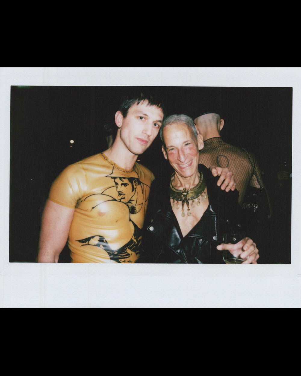 Two men posing together, one wearing a yellow printed shirt and the other in a black leather outfit holding a drink.