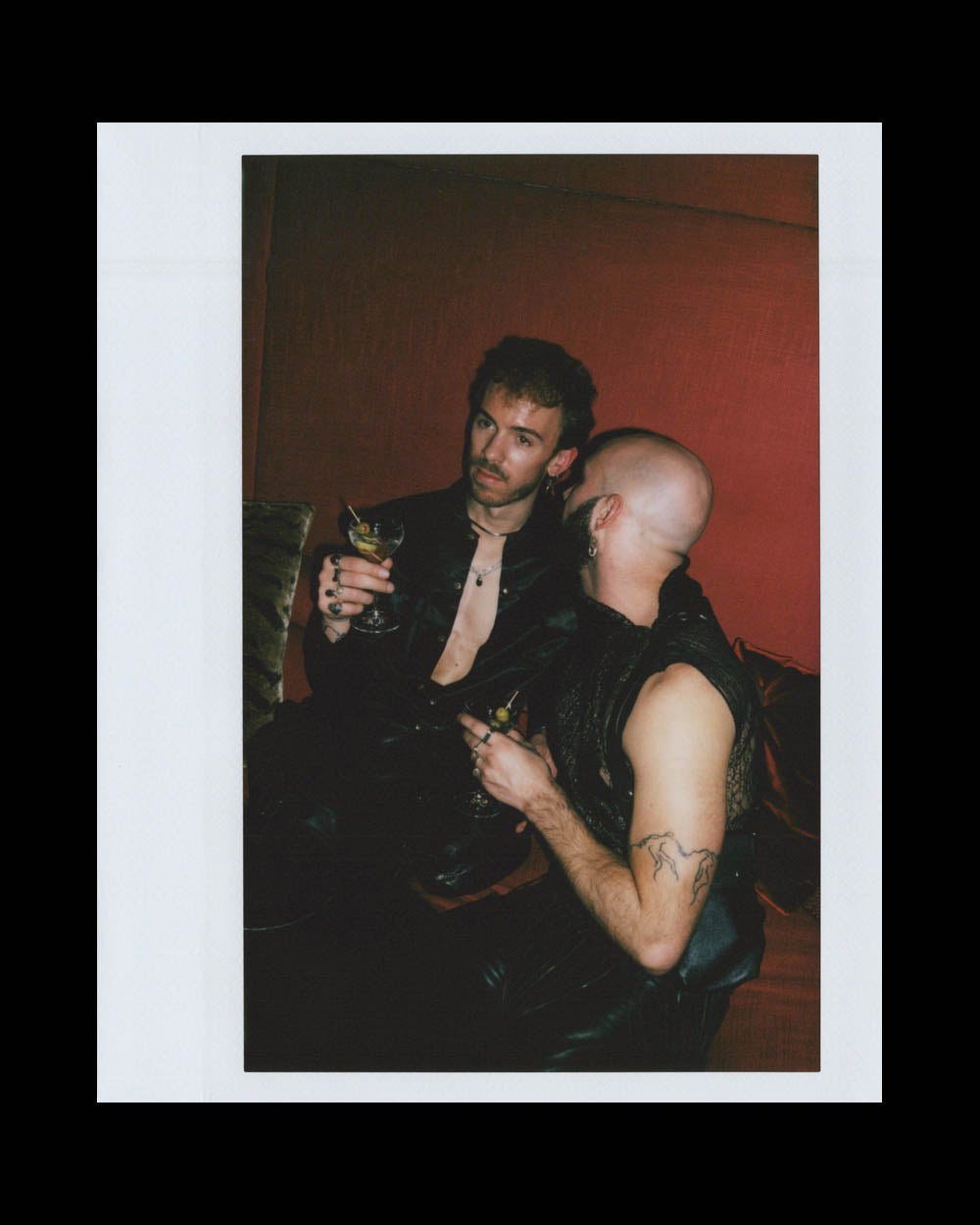 Two men dressed in black leather seated closely on a red couch, each holding a cocktail glass.