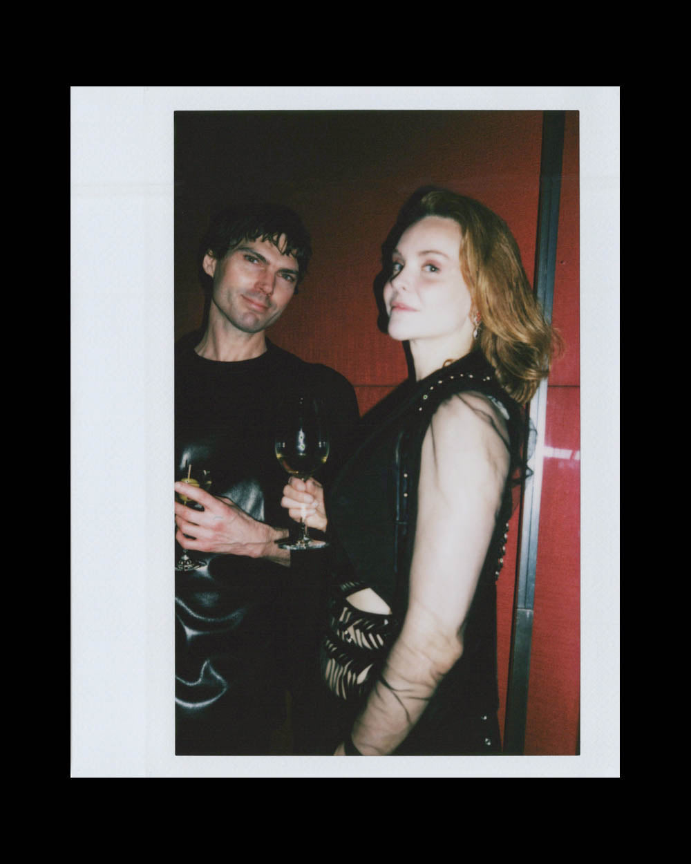 Man and woman holding wine glasses standing against a red wall, both dressed in black evening attire.
