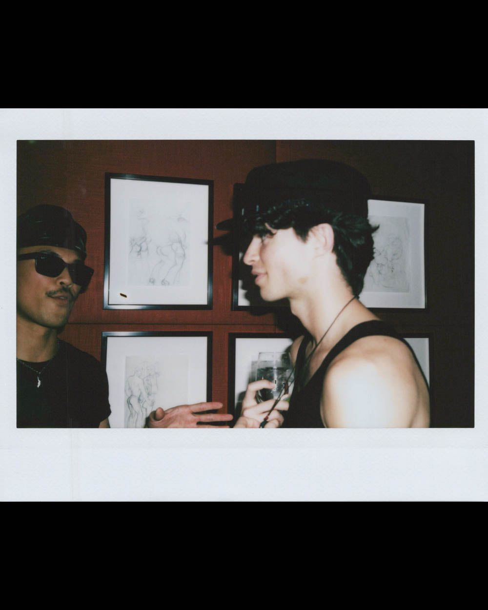 Two men wearing hats talking in front of framed sketches on a wooden wall indoors.