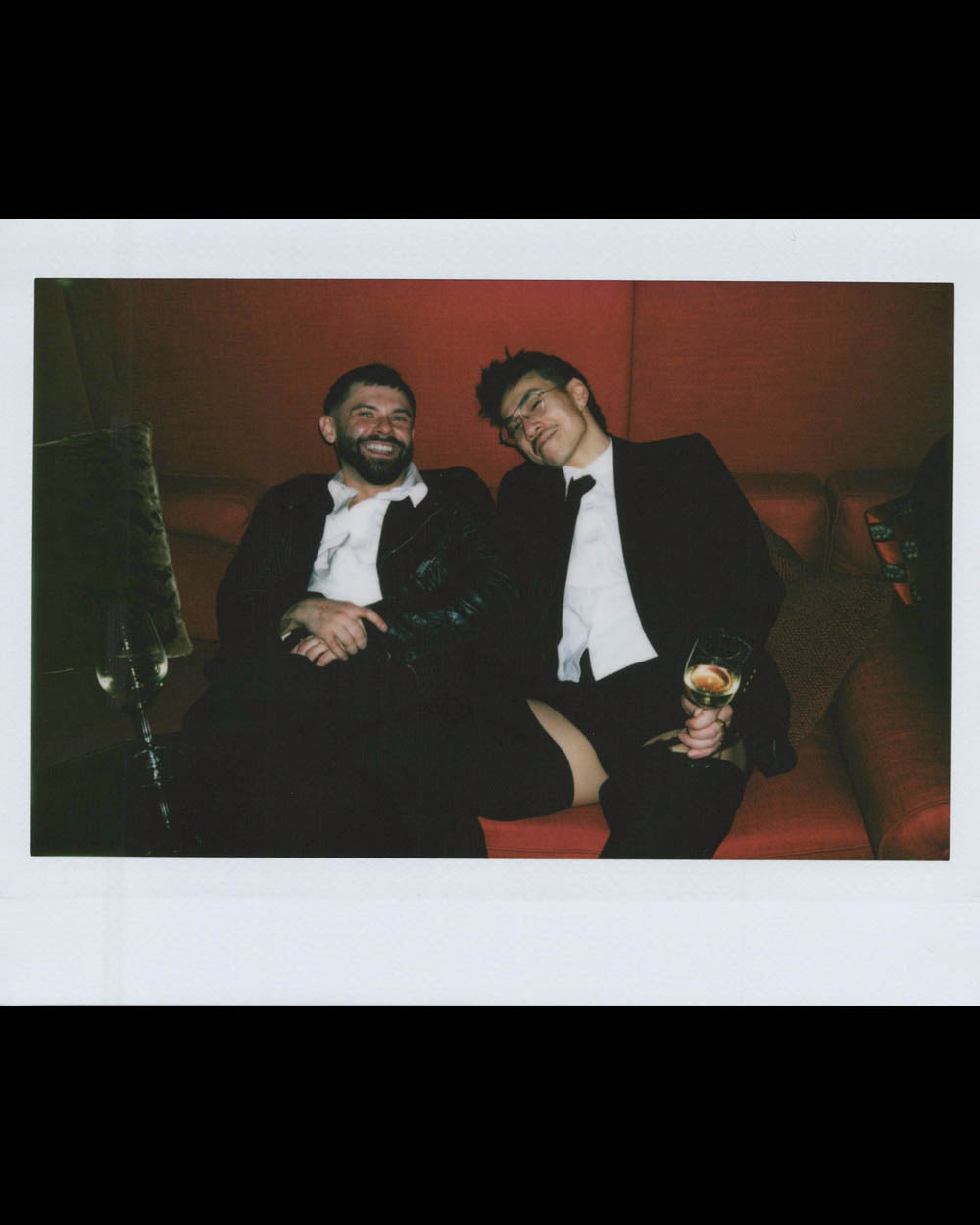 Two men in formal attire reclining on a red sofa, one holding a glass of champagne with a relaxed expression