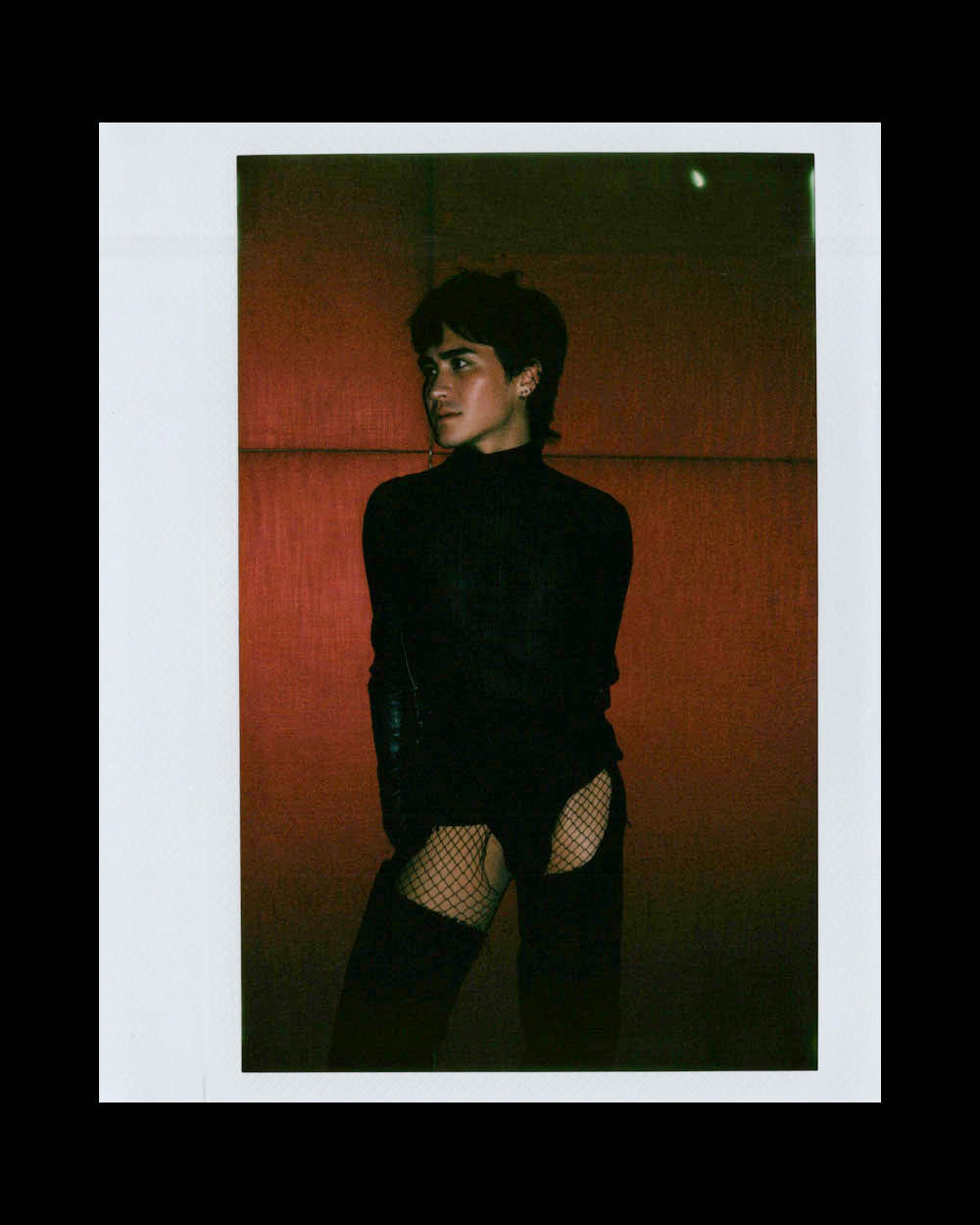Person in a black long-sleeve top and fishnet stockings posing against a deep red padded background