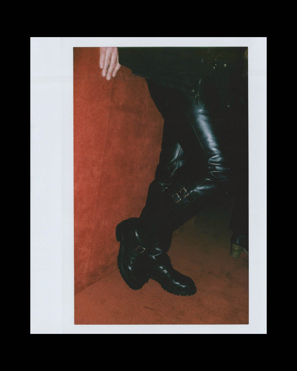 Person wearing black shiny leather boots leaning against a burnt orange textured wall with one hand resting above.