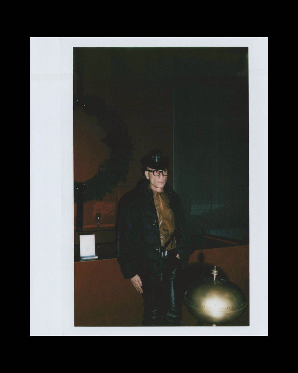 Person wearing a black jacket, leather pants, and a cap standing indoors next to a brass decorative object.