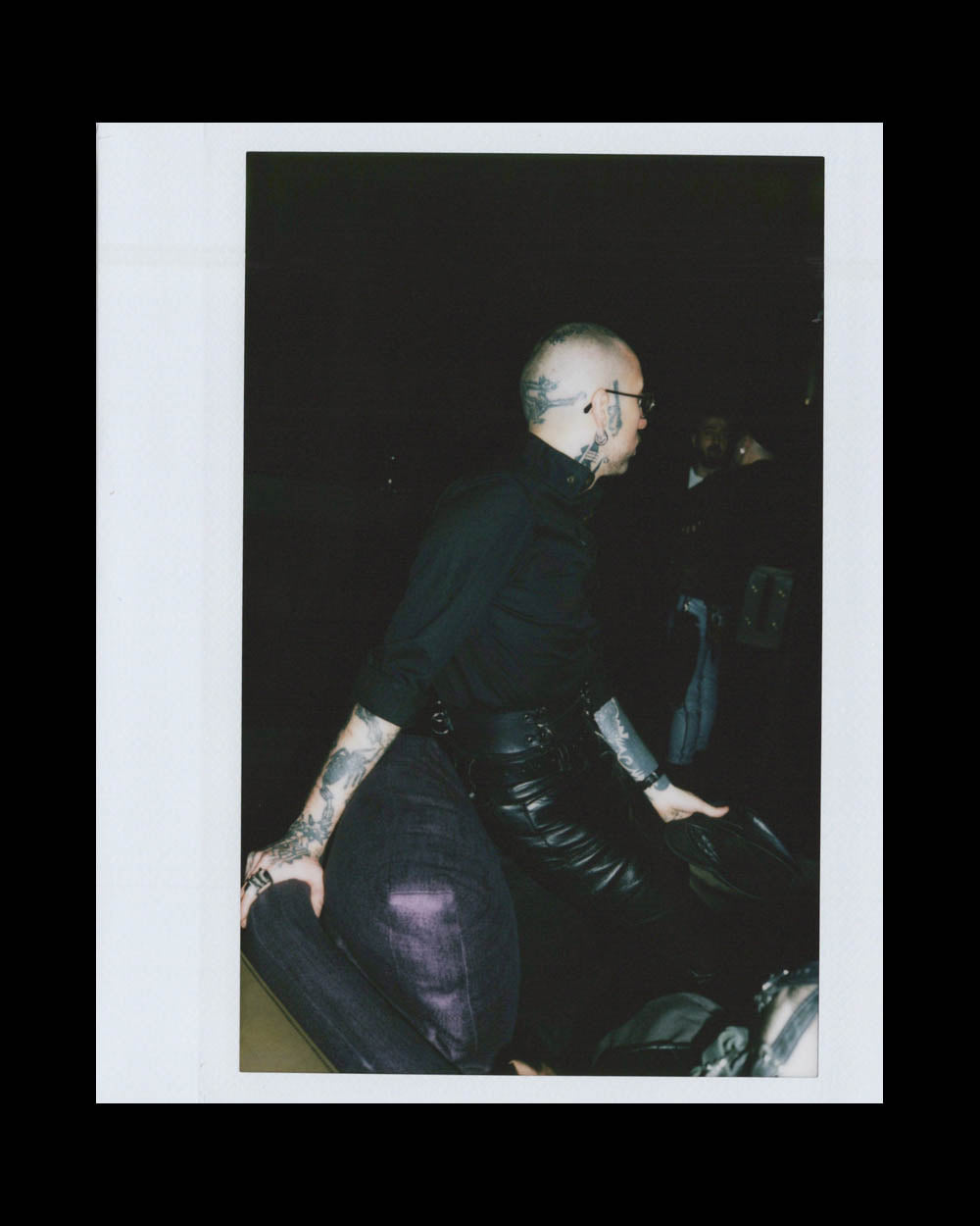 Person with tattoos and glasses wearing black leather pants seated on a dark cushioned chair in a dim setting.