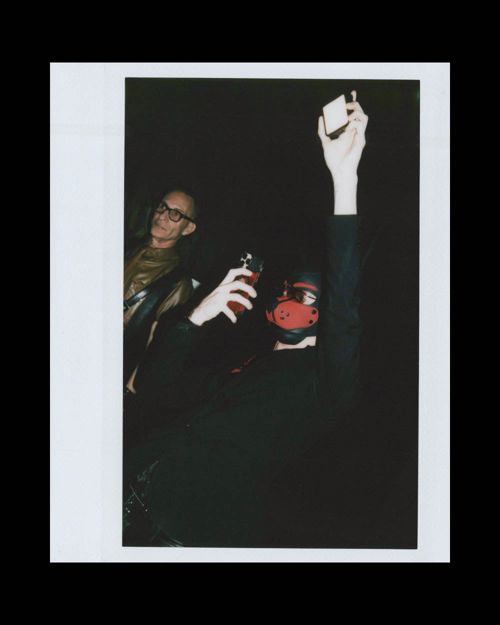 Person wearing a red mask and dark clothing raising a phone while another person with glasses and a leather jacket stands nearby in dim light.