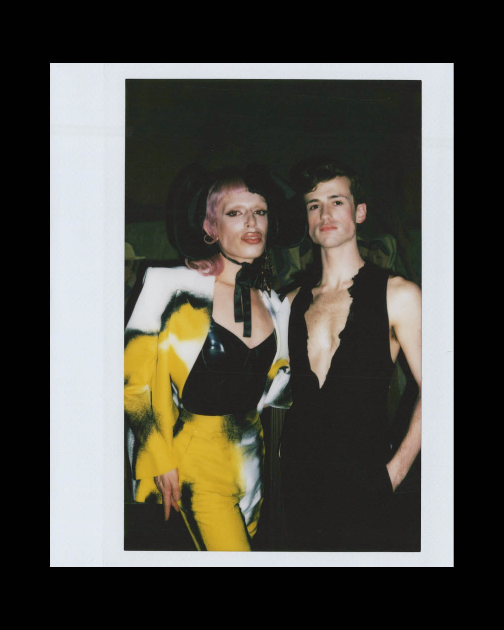 Two individuals posing; one wearing a yellow and black patterned suit with a wide-brimmed hat, the other in a sleeveless black top.