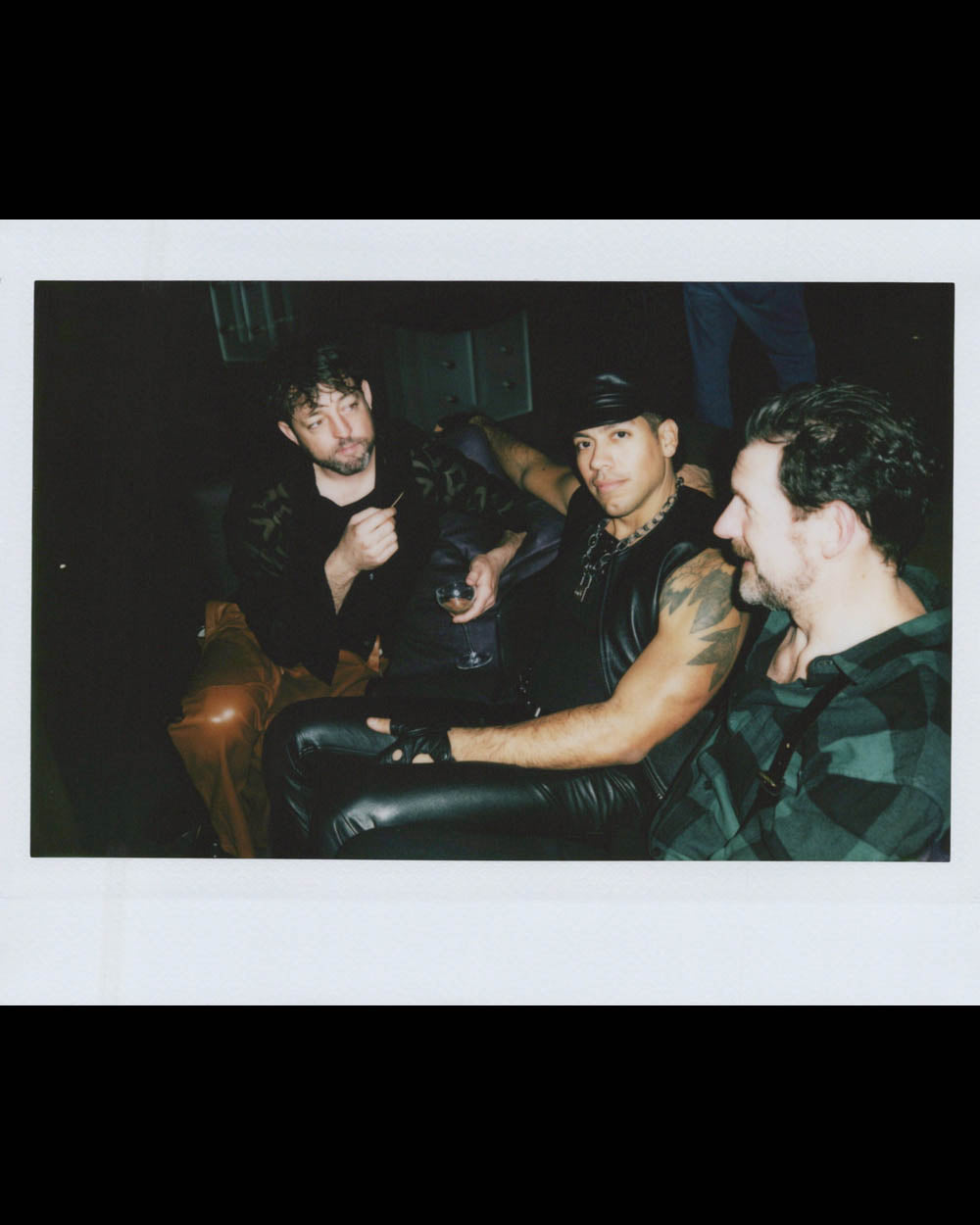 Three men sitting closely together, one wearing leather and a cap, another in casual plaid shirt, in a dimly lit space.