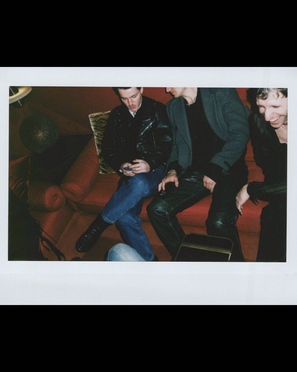 Three people sitting on a brown couch, two wearing black leather clothing, one smiling and leaning forward.