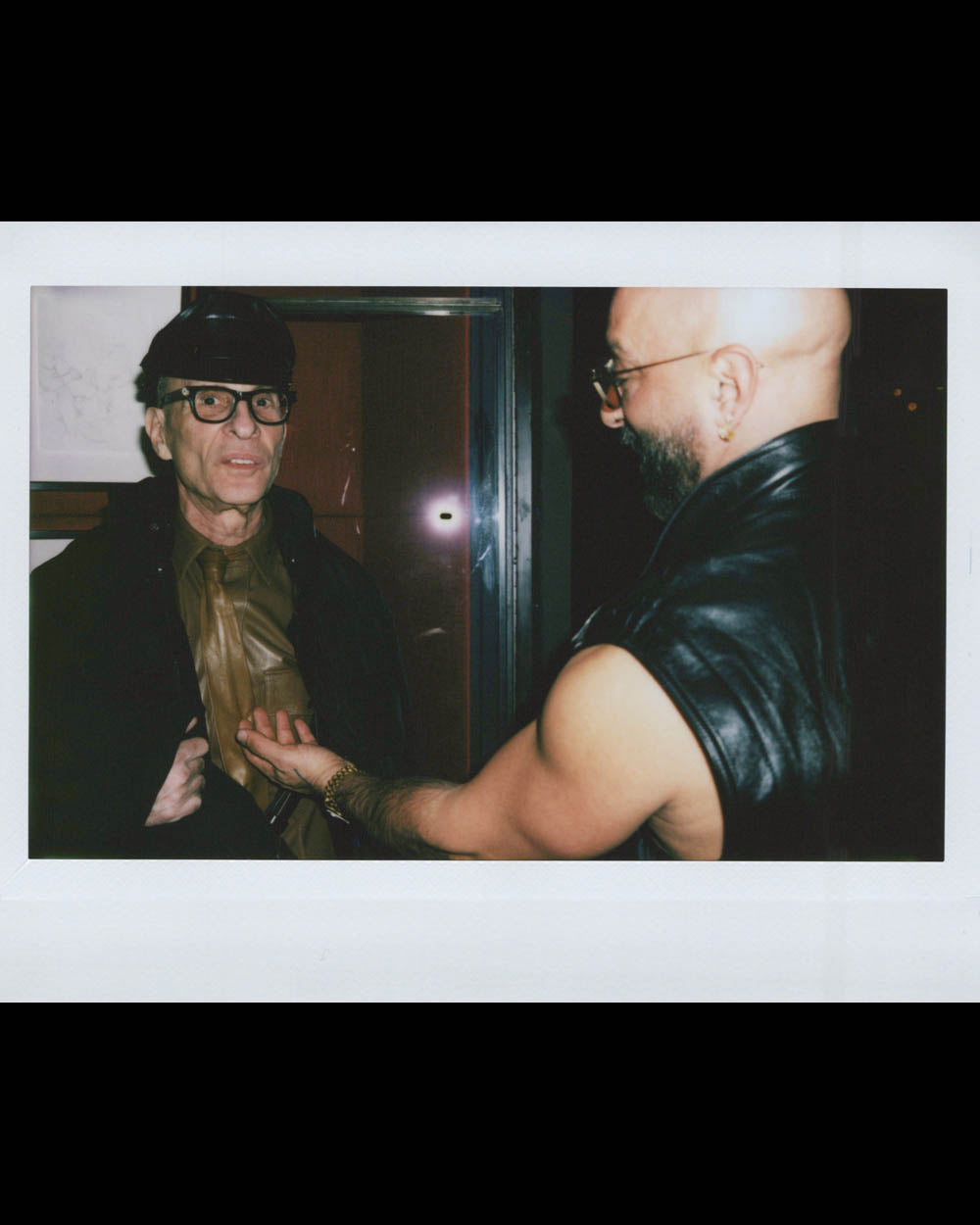 Two men in leather clothing engaged in conversation, one wearing glasses and a sleeveless jacket indoors.