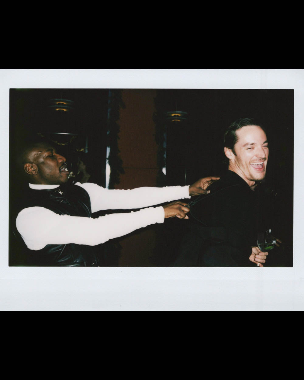 Two men laughing together, one reaching out to touch the other’s shoulders in a dark indoor setting.