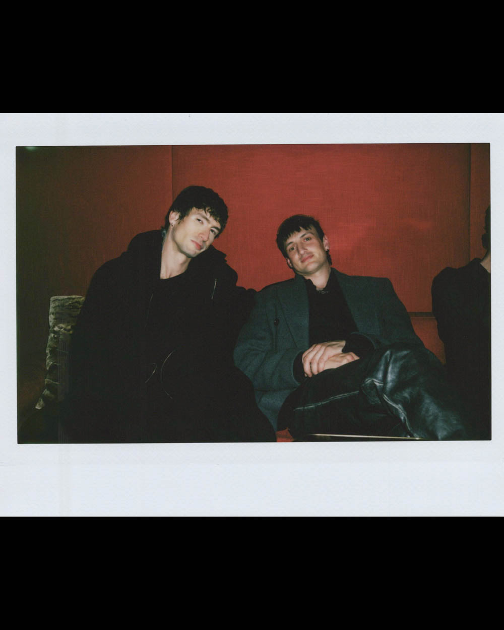 Two men sitting side by side on a couch against a red wall in a relaxed indoor setting.