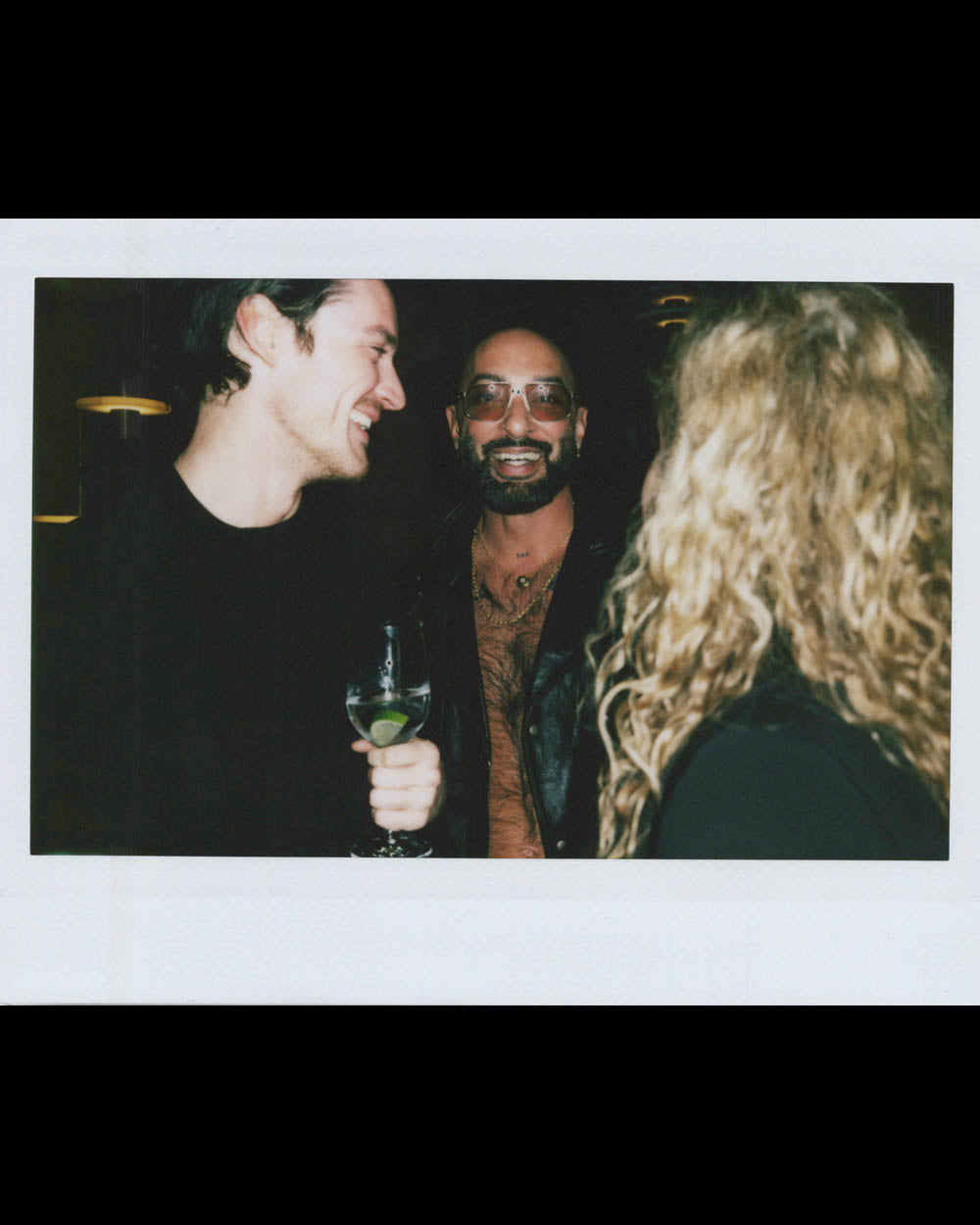 Three people socializing indoors, one holding a drink, another wearing sunglasses and leather clothing.