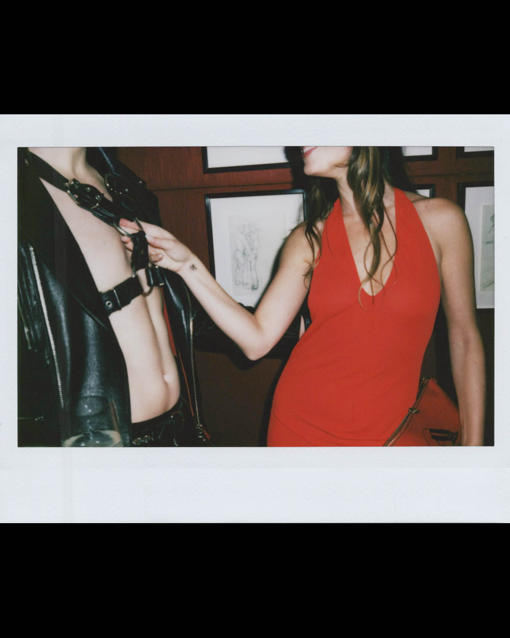 Woman in a red dress touching a person wearing a black leather harness and jacket indoors against a framed wall background.