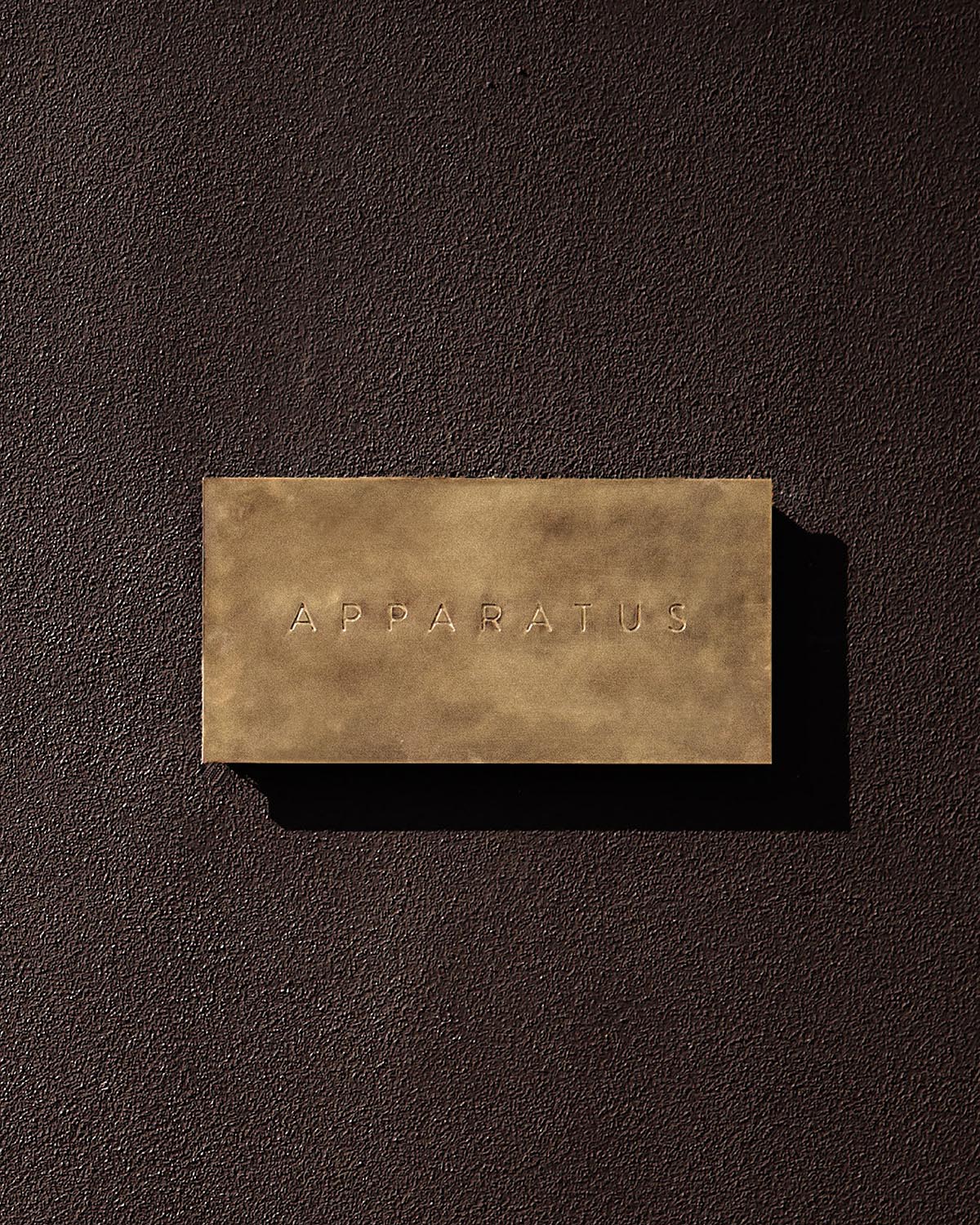 Rectangular tan leather tag embossed with the word "APPARATUS" casting a shadow on a textured dark surface.