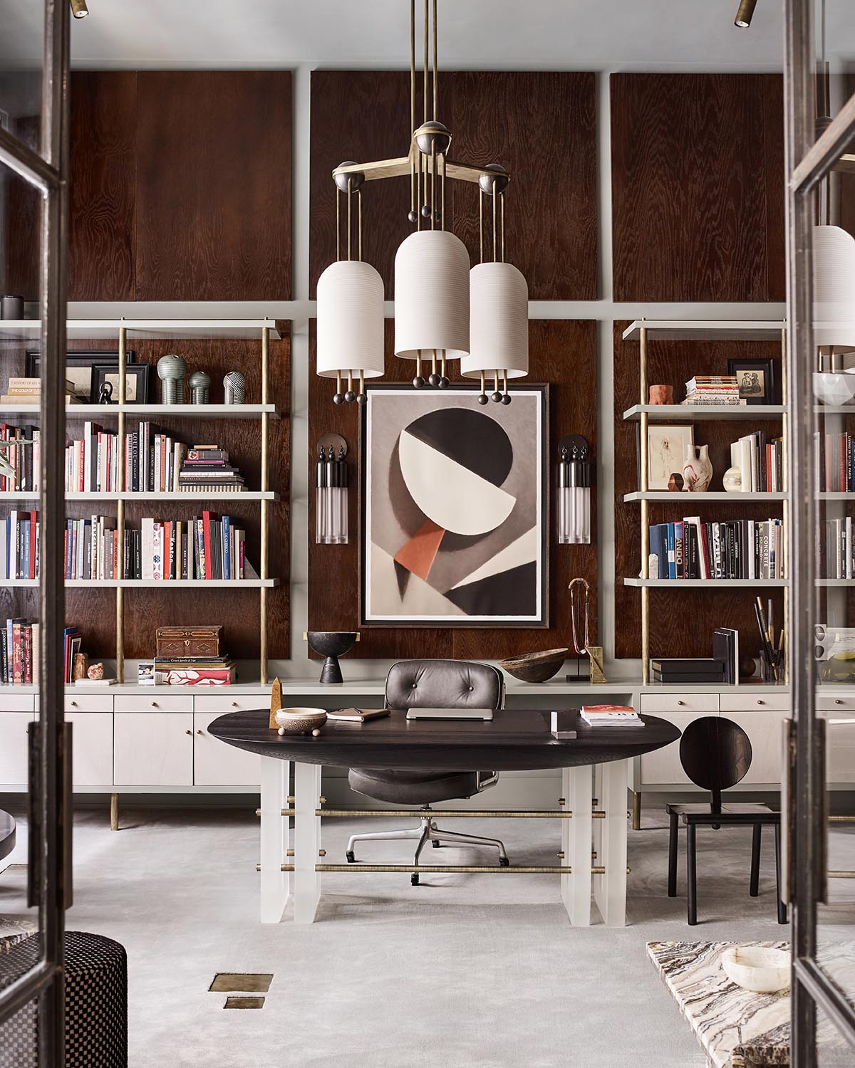 Modern office space with dark wood paneled walls, geometric artwork, oval black desk, and open shelving units