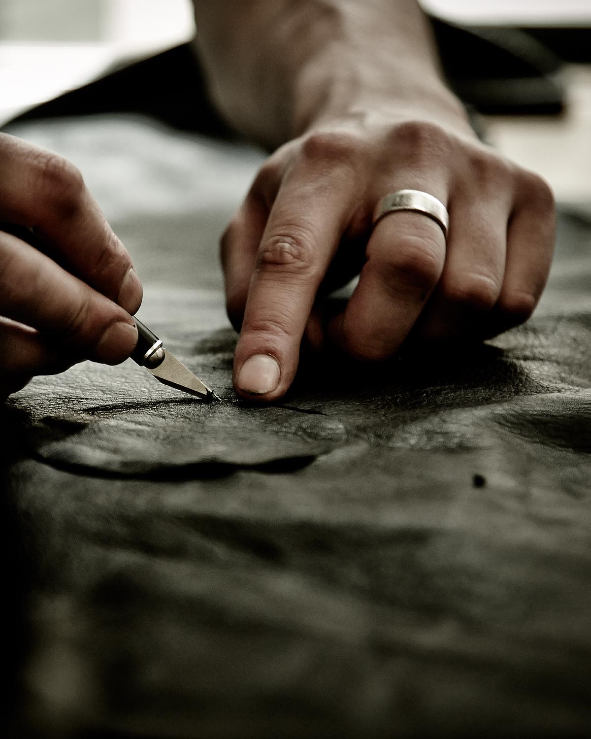 Close-up of hands carving or cutting textured material with a small blade, one hand wearing a silver ring, on a dark surface