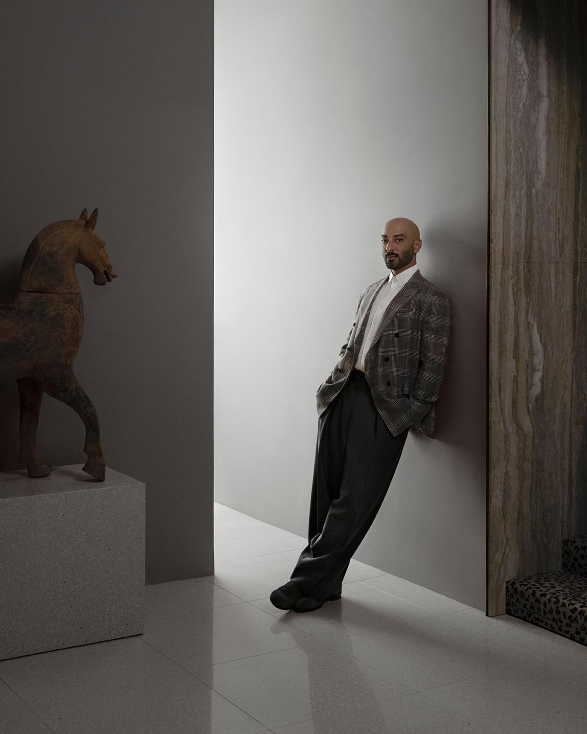 Man in plaid blazer and dark trousers leaning against a light gray wall in a minimalist interior space