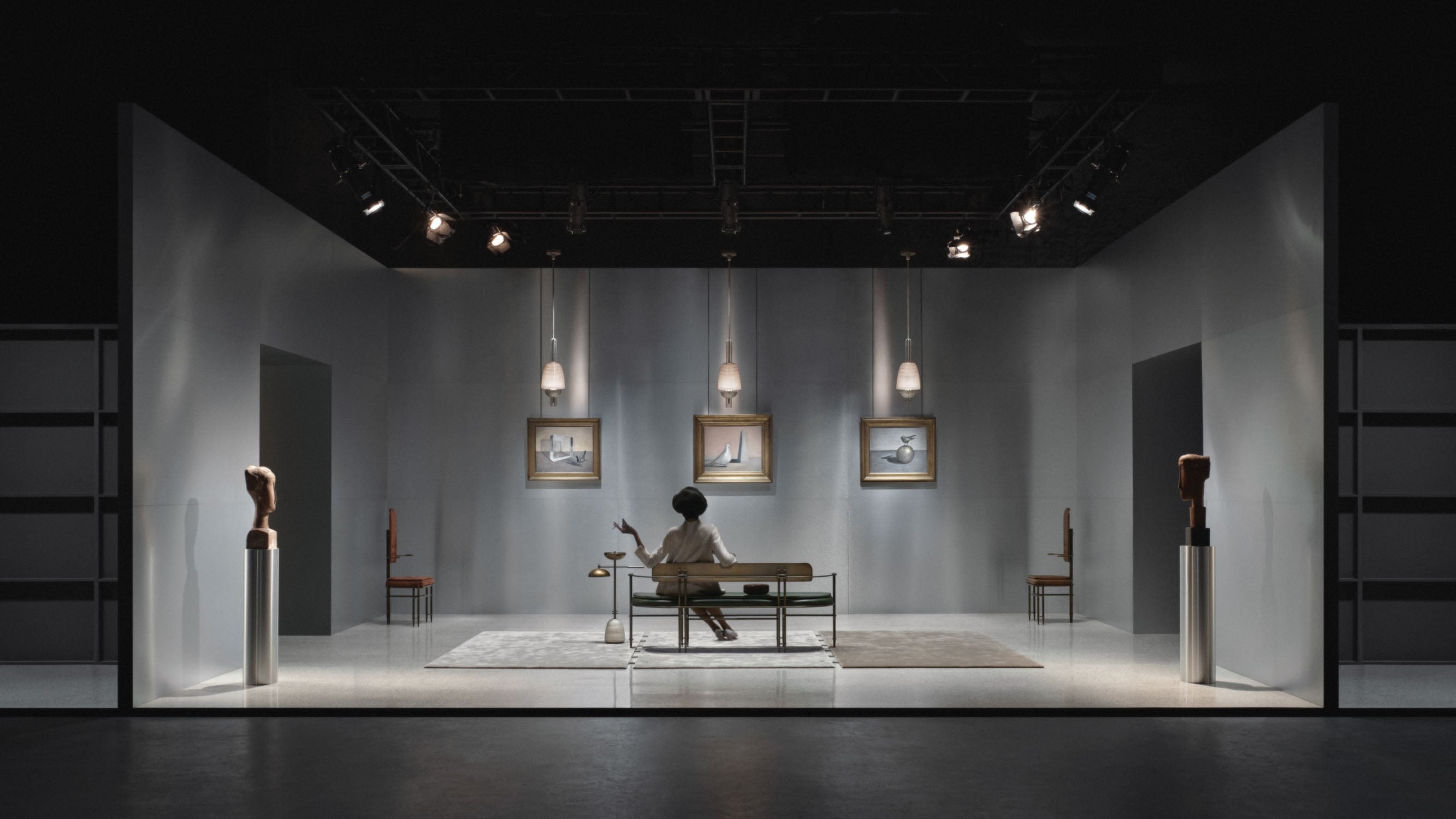 Theatrical set featuring a person sitting on a bench surrounded by framed sketches, pendant lights, and sculptural pieces on pedestals.