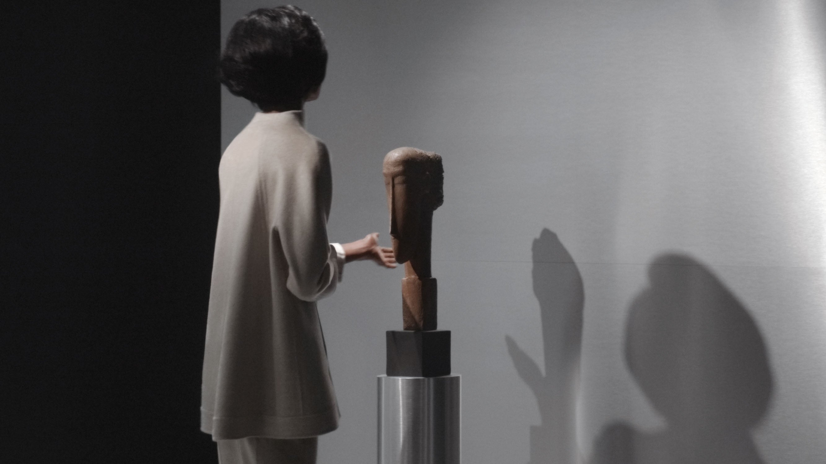 Person with short dark hair standing beside a wooden abstract sculpture casting a shadow on a plain wall