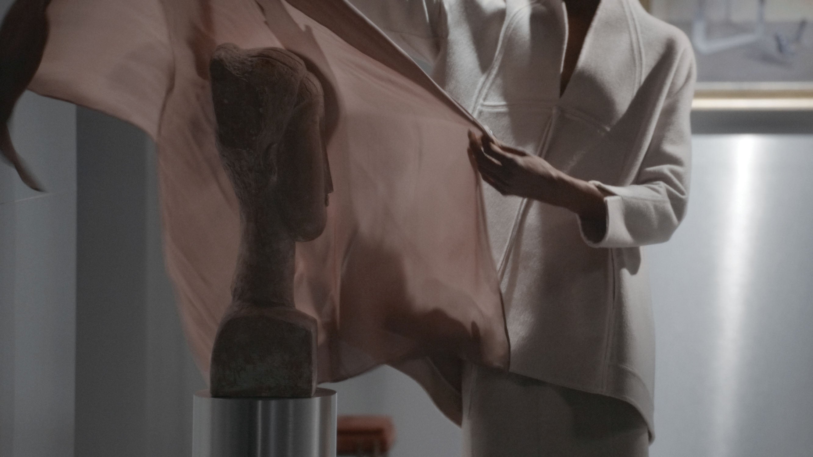 Person draping a sheer fabric over a stone bust sculpture in a softly lit interior setting