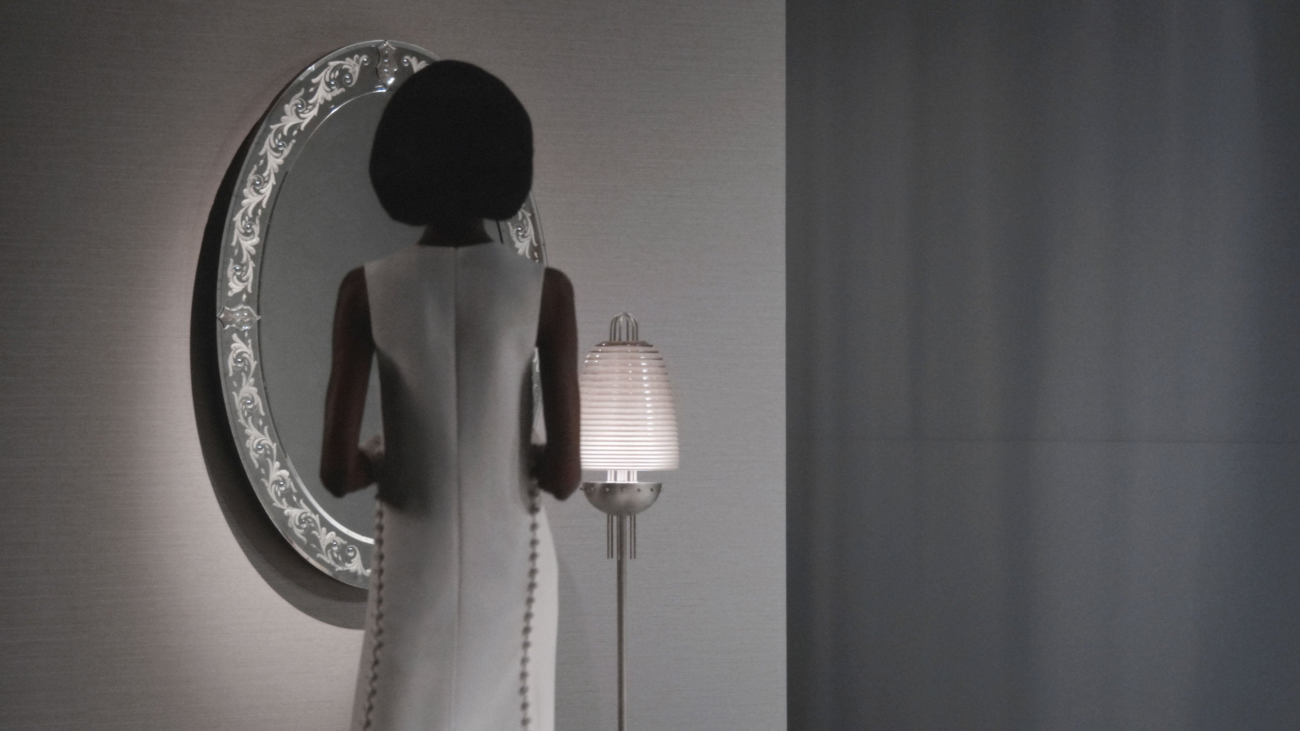 Woman in a white sleeveless dress standing in front of an ornate oval mirror beside a floor lamp with a textured shade