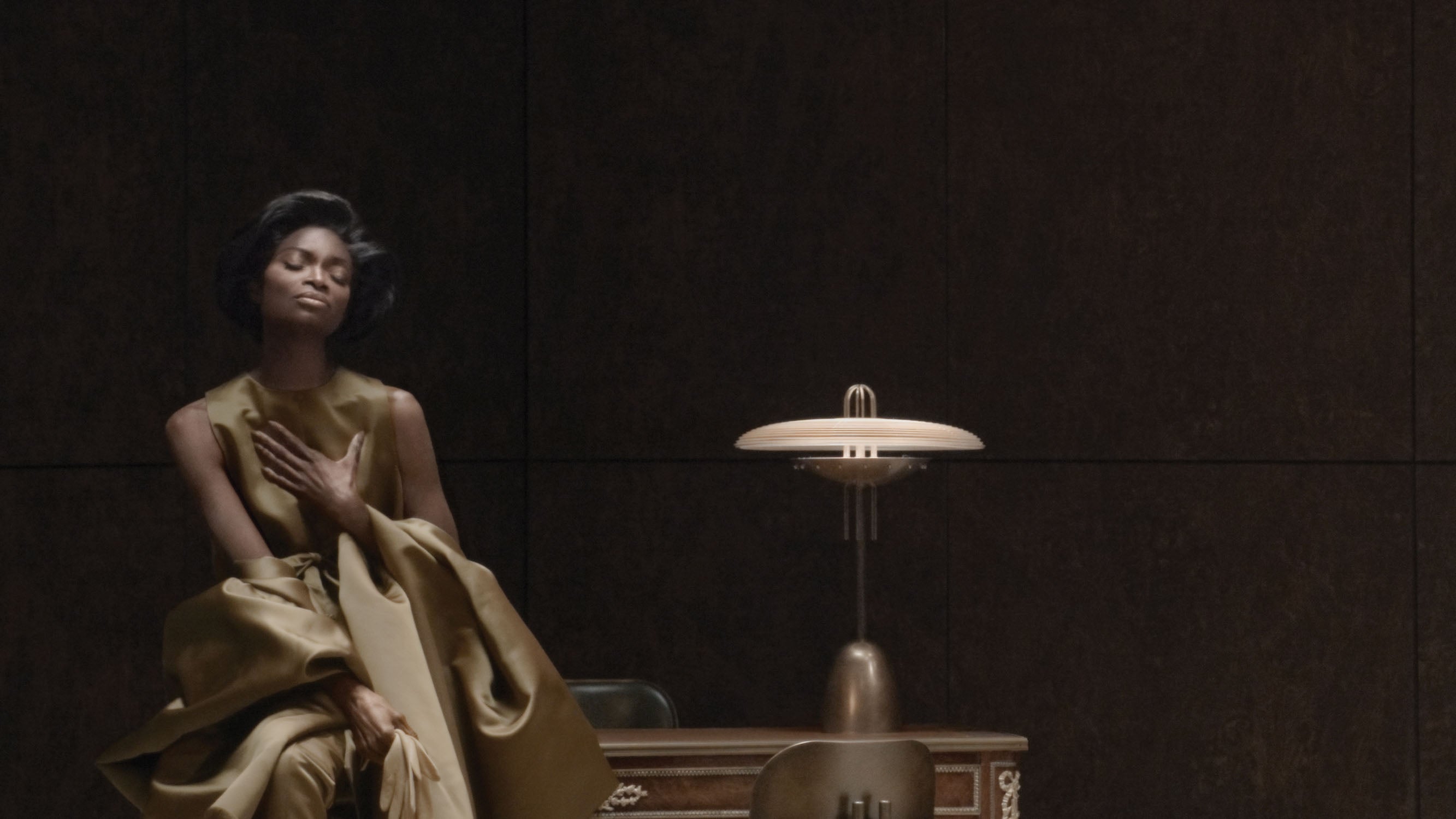 Woman in textured gold fabric dress sitting beside a vintage table lamp against a dark brown wall panel background
