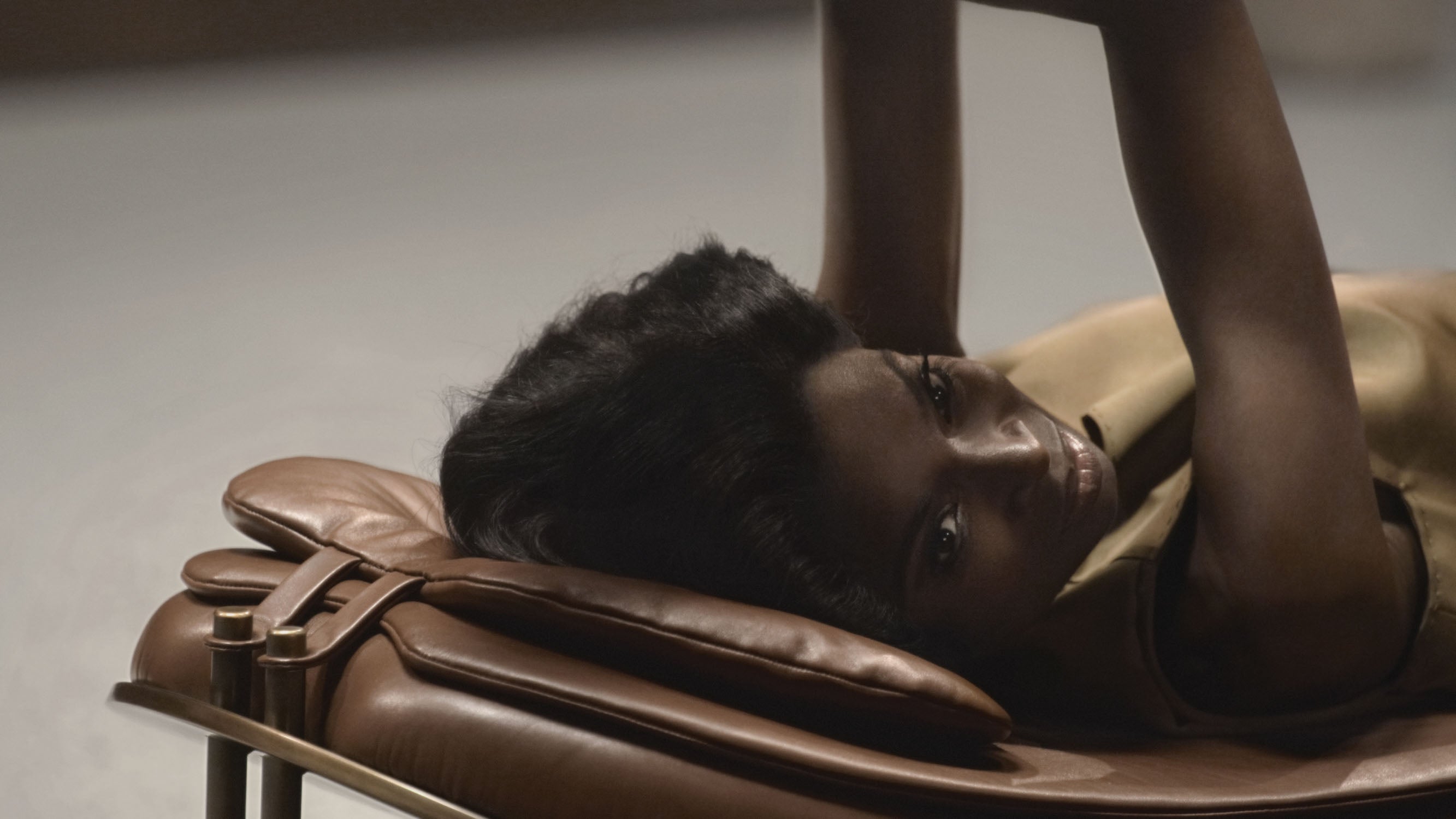Side view of a woman lying on a brown leather chair with her arm raised against a neutral background