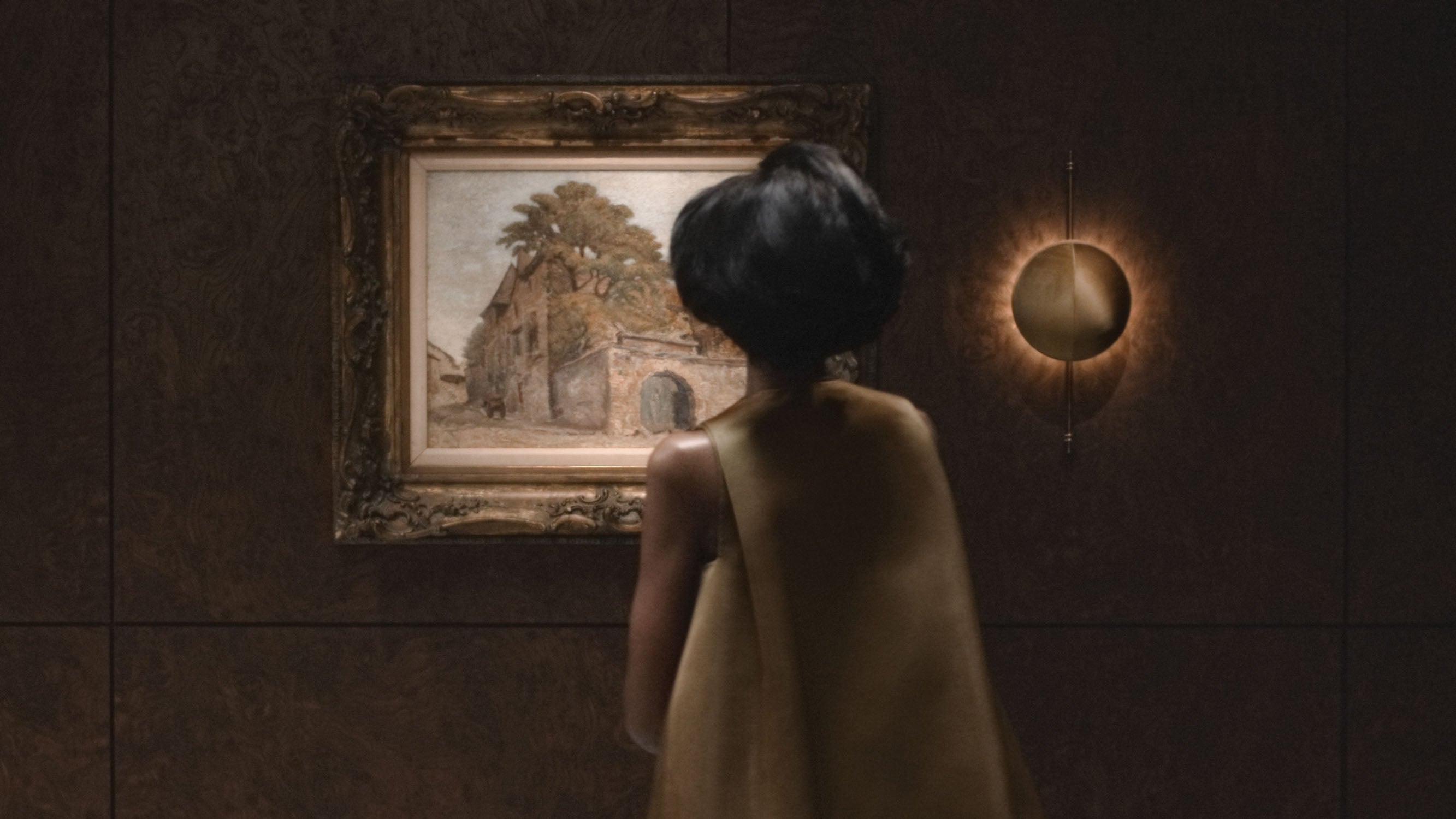 Woman with short dark hair wearing a sleeveless beige dress looking at framed landscape painting on dark wall