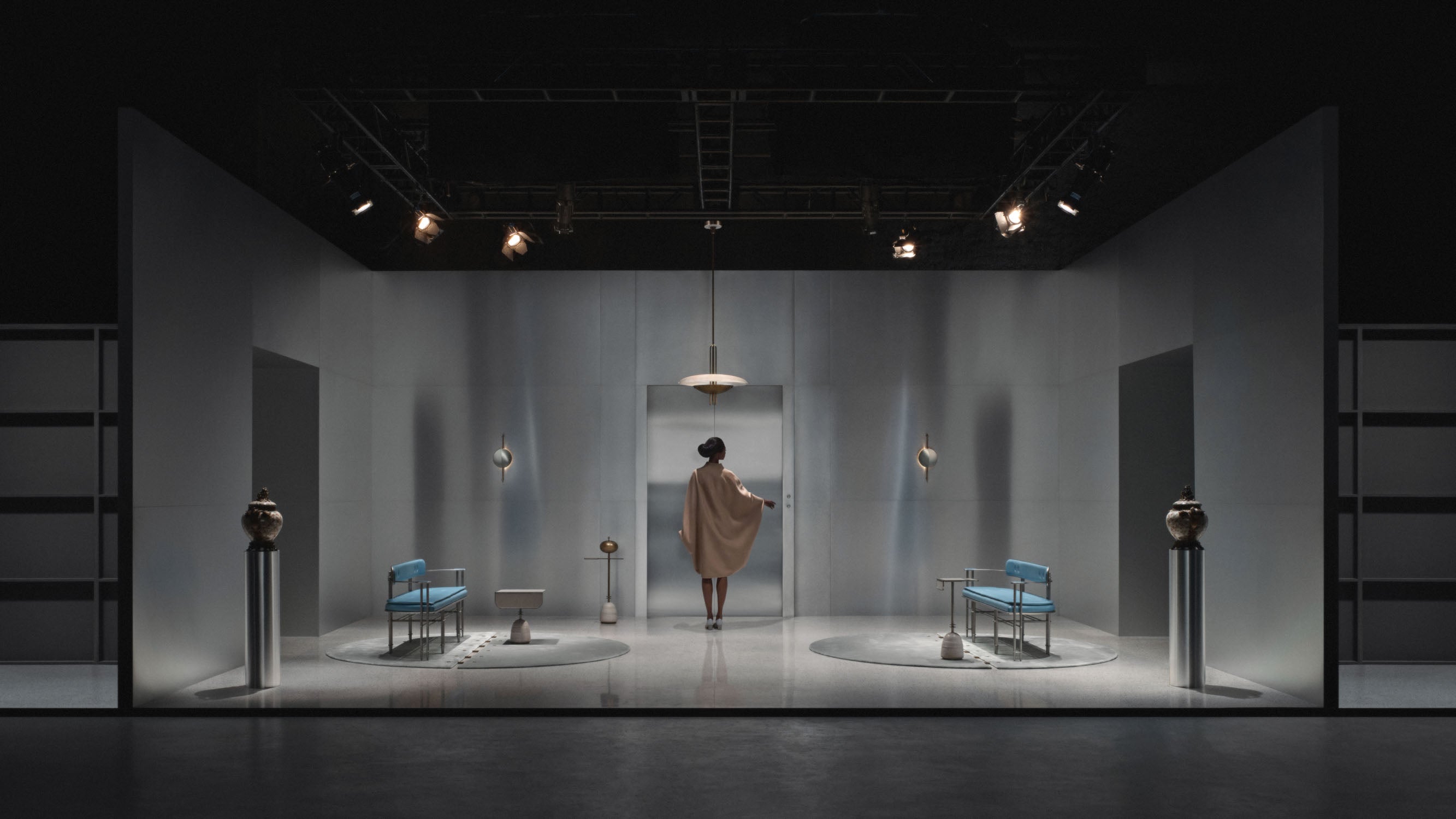 Wide view of a modern minimalist stage set with a person in a beige coat facing a door in the center under soft lighting