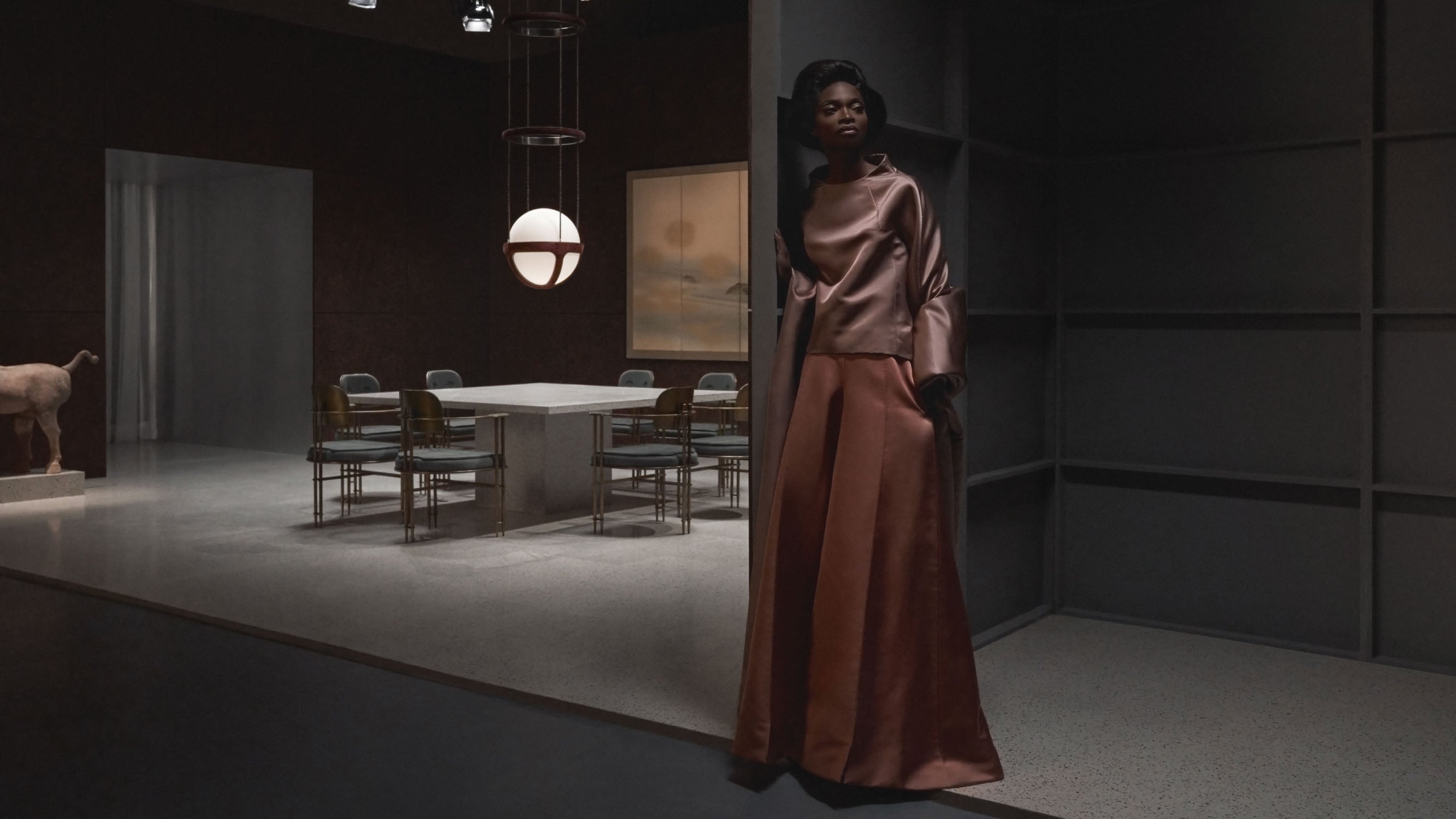Woman in a bronze satin dress standing in a dimly lit modern room with dining table and artwork in the background