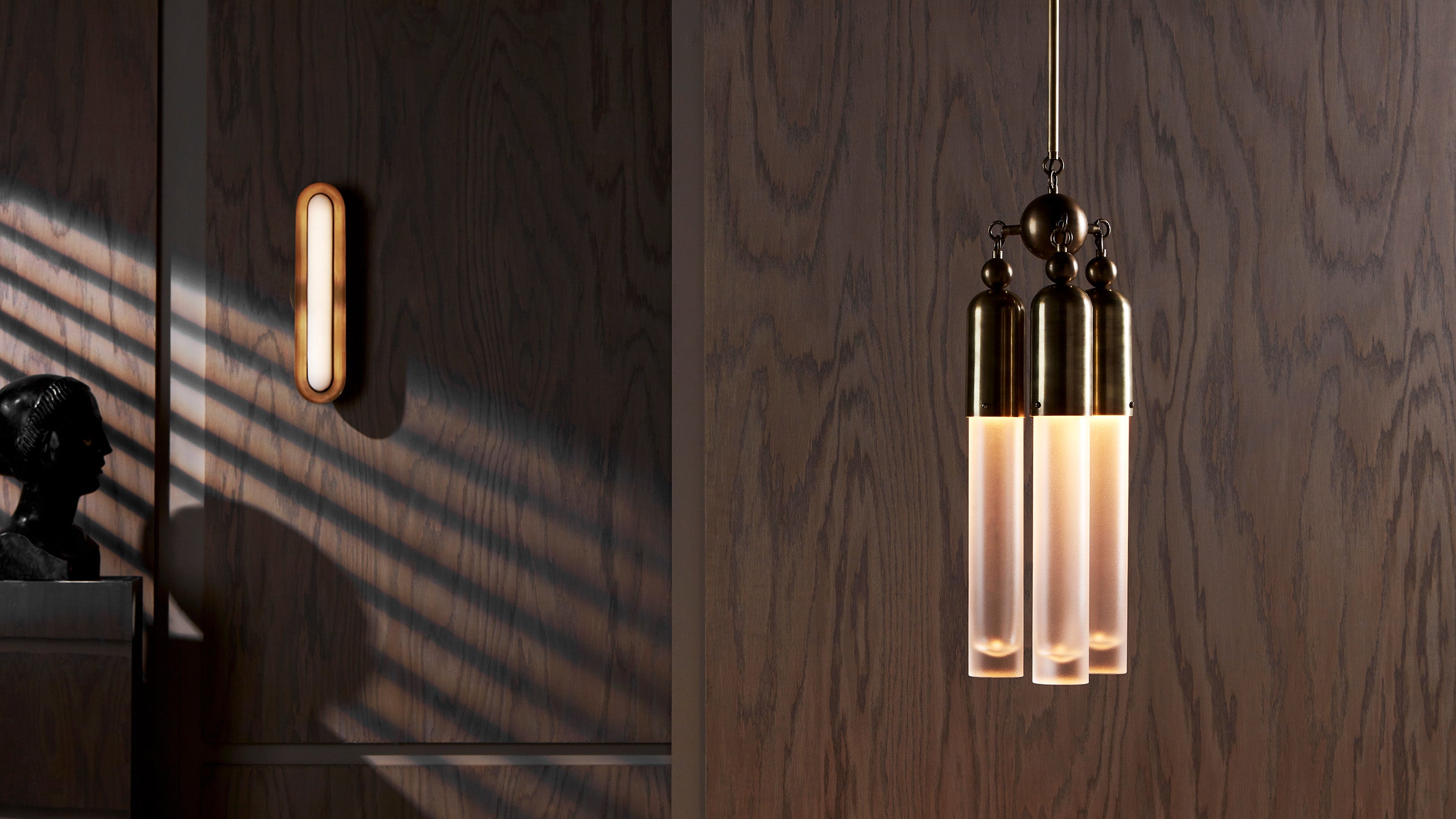 Brass pendant light with frosted glass tubes hanging against a dark wood paneled wall with soft window light shadows