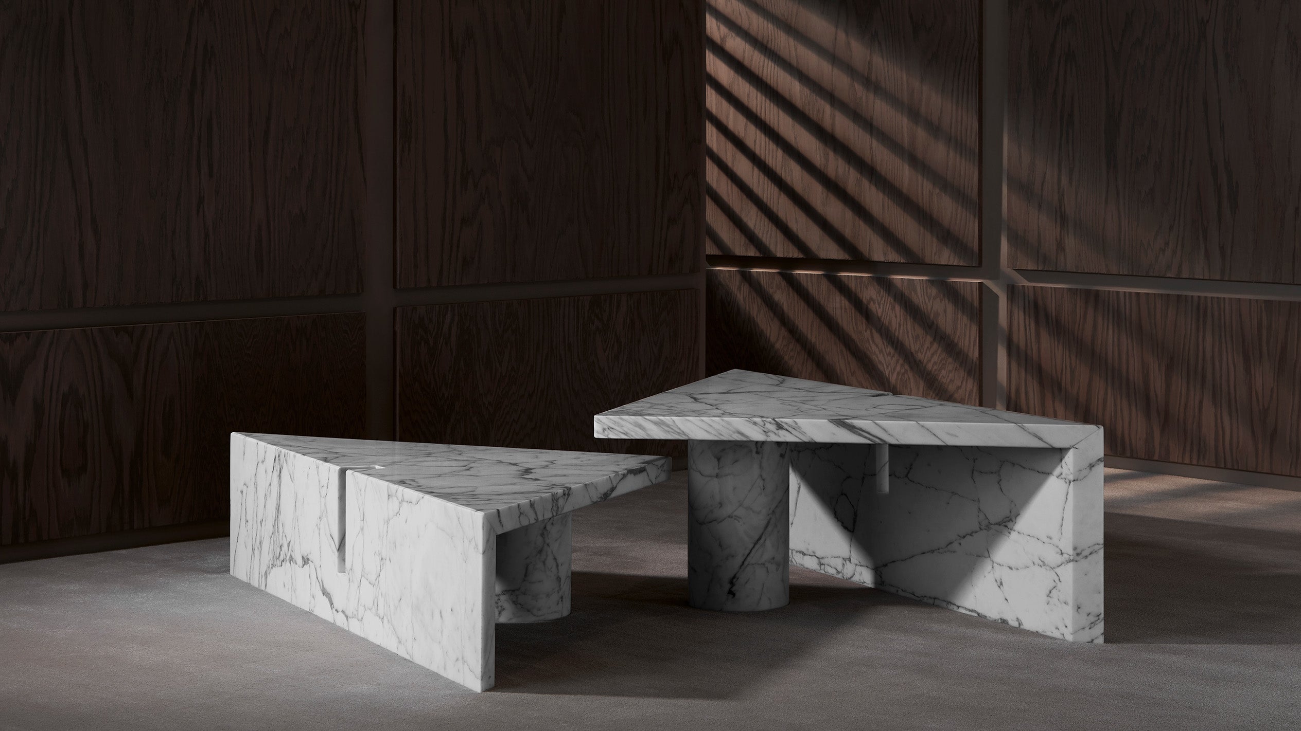 Two triangular white marble tables with gray veining on a concrete floor against dark wood panel walls with sunlight shadows