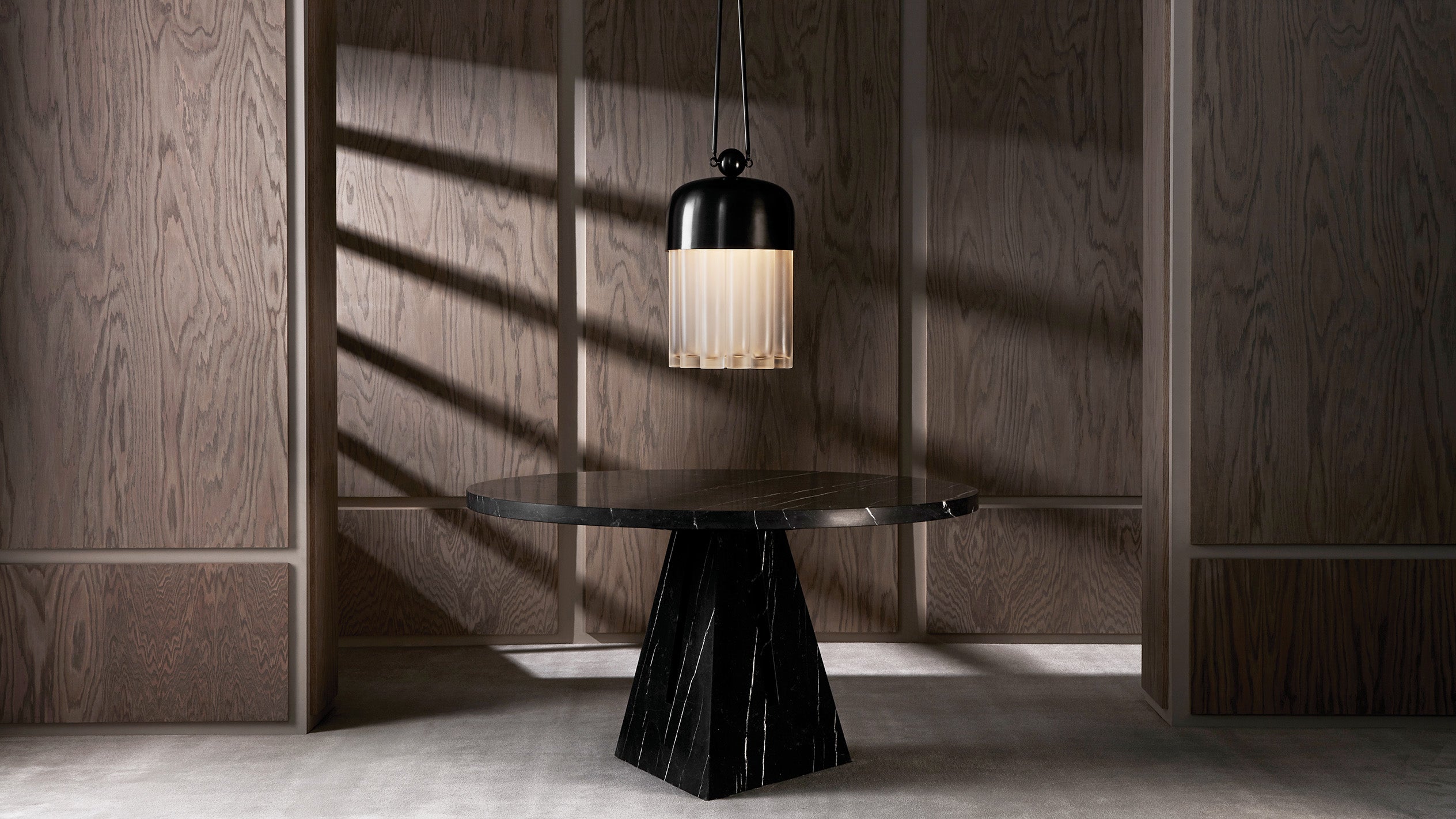Round black marble table with geometric base under a suspended black and white pendant light in wood-paneled room