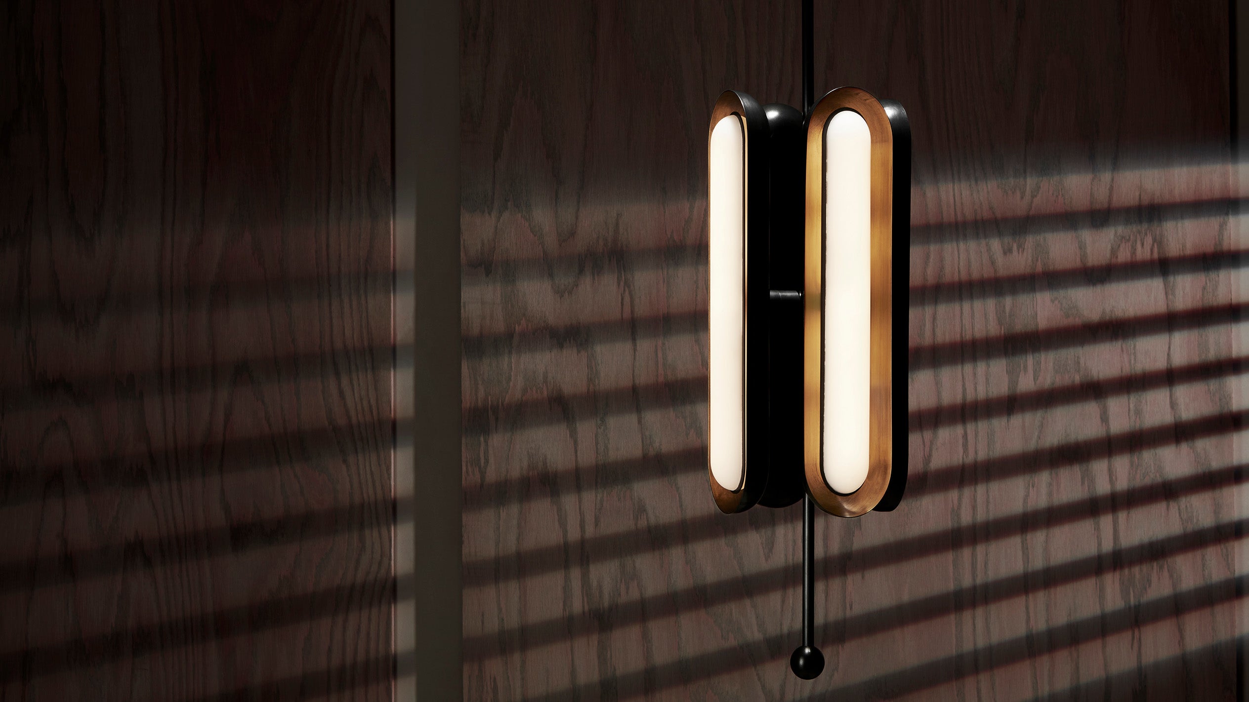 Vertical oval-shaped wall light with warm glow casting striped shadows on a textured dark wooden wall
