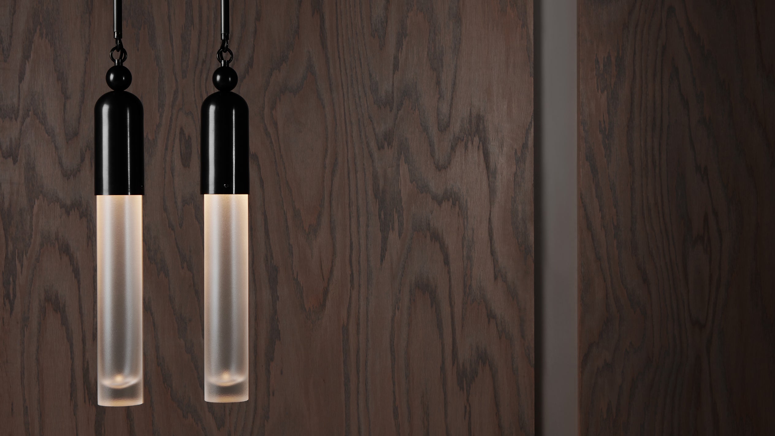 Two slender cylindrical pendant lights with black tops and frosted glass hanging against a dark wood panel wall