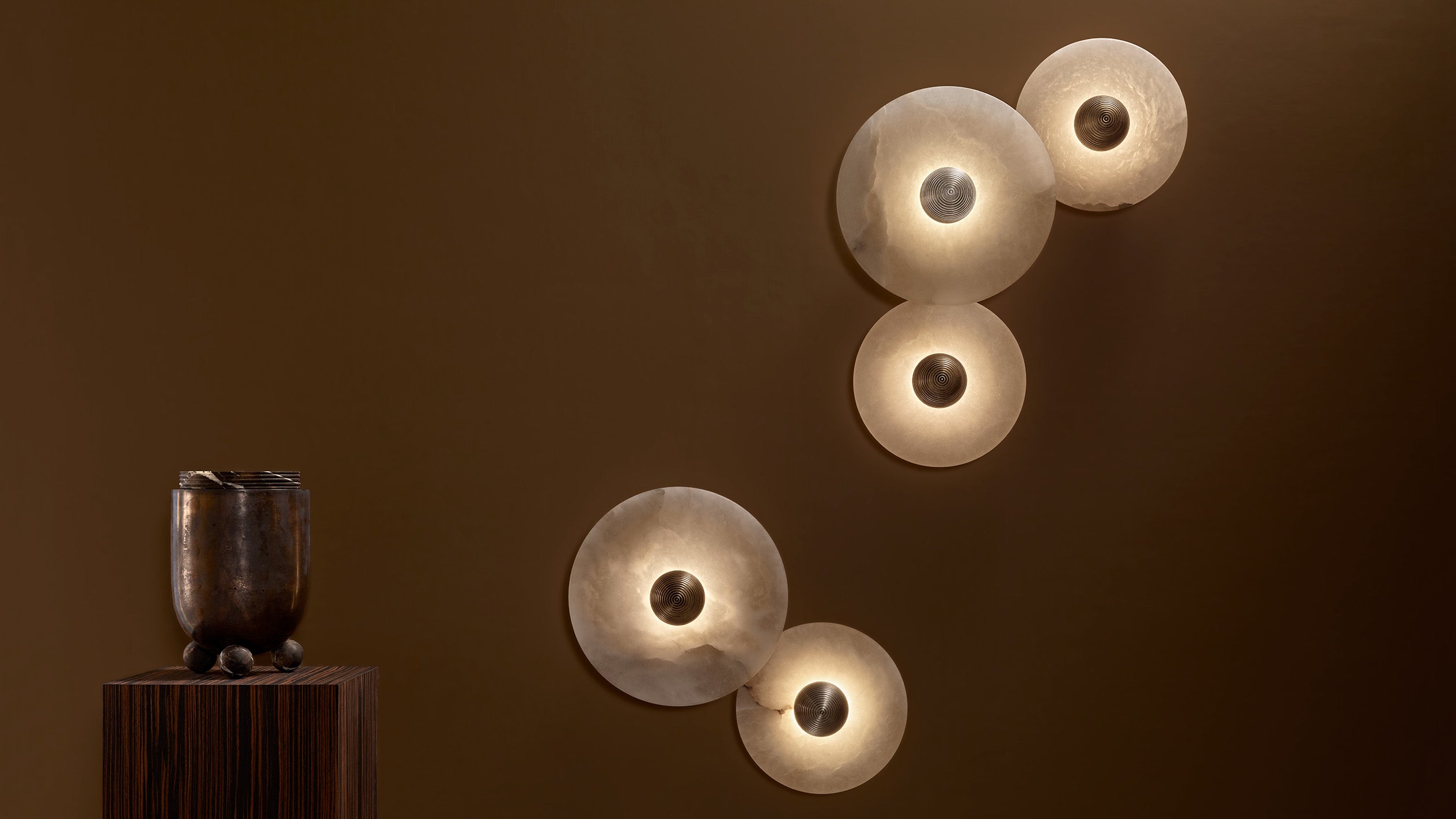 Warm ambient light from four circular wall lamps arranged asymmetrically on a smooth brown wall above a dark wooden pedestal with a sculptural vase