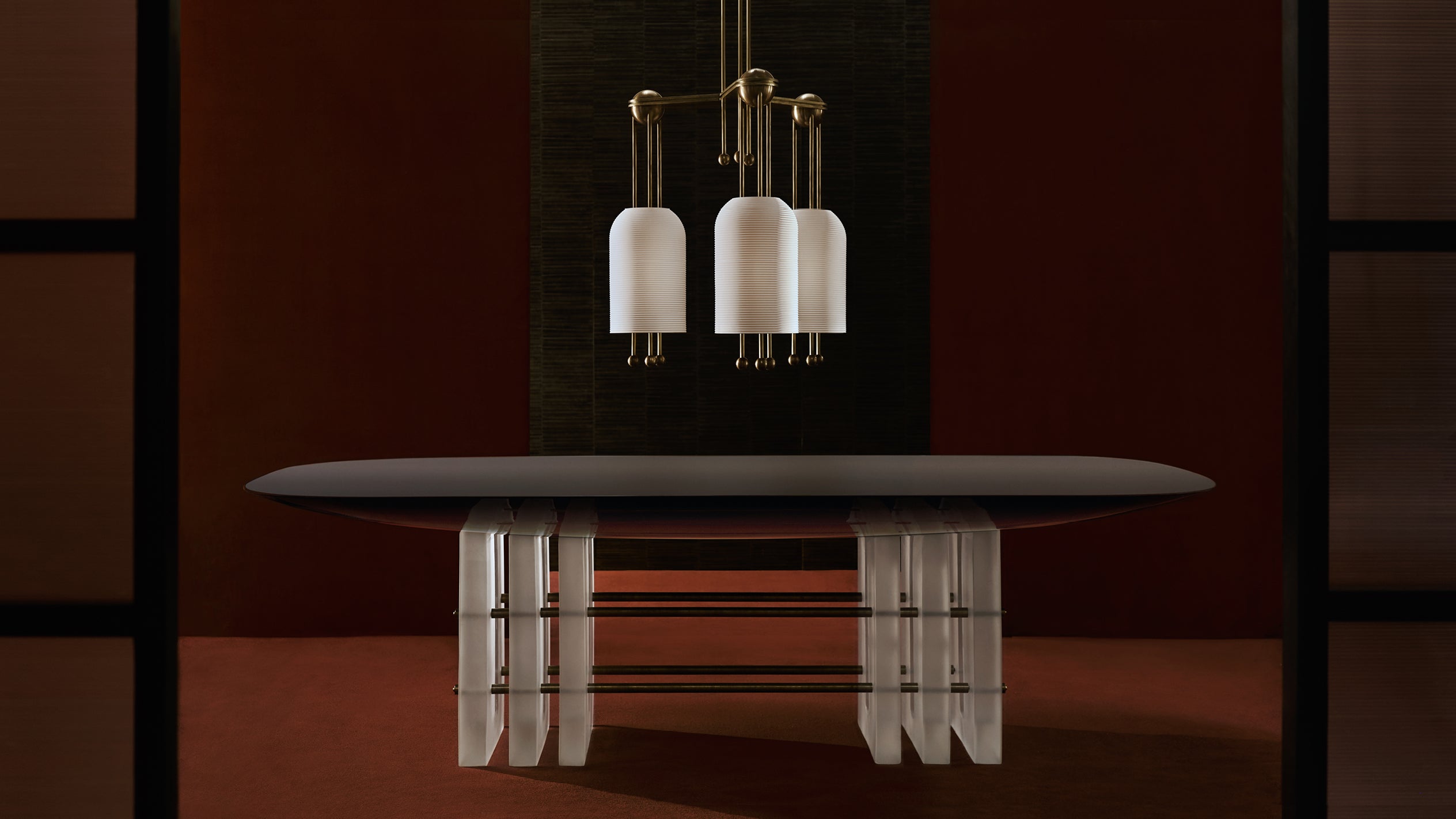 Oval dark tabletop supported by translucent sculptural legs under pendant lights with white cylindrical shades against a dark backdrop