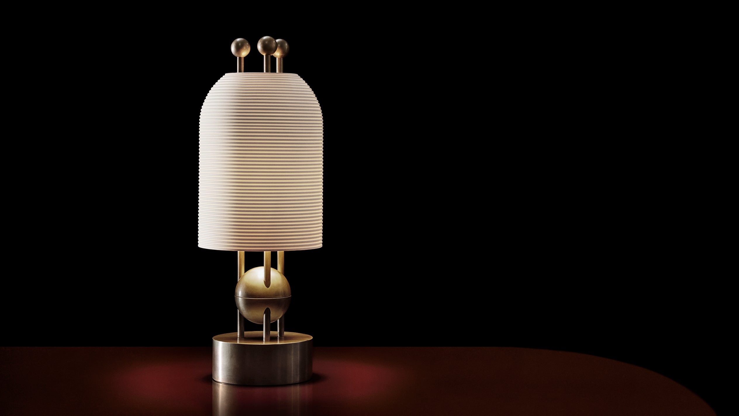 Modern table lamp with a ribbed cream shade, brass spherical accents, and a cylindrical metal base on a dark background