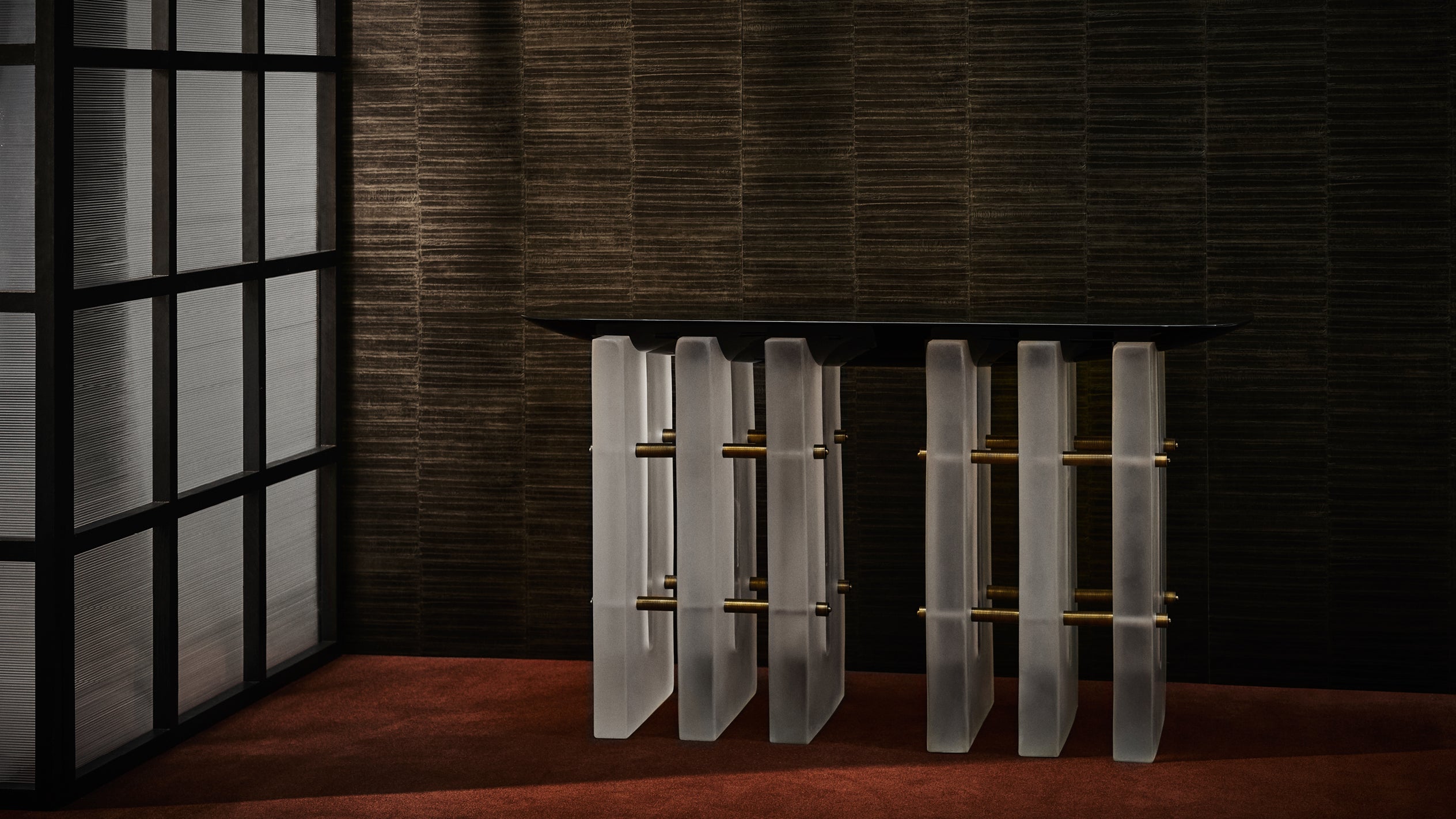 Side view of a modern table with vertical frosted glass panels and brass rods against a textured brown wall