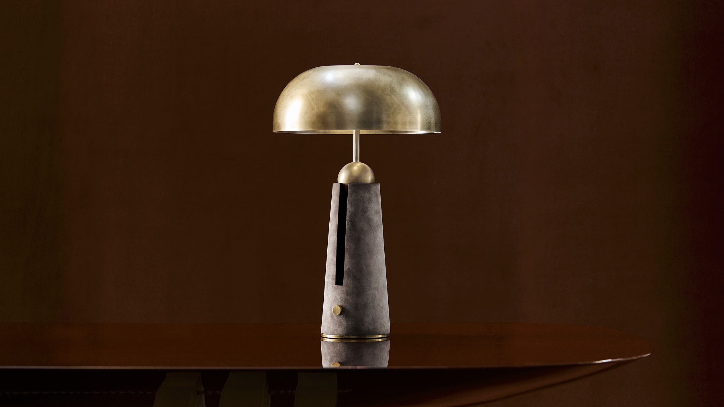 Sculptural table lamp with brass dome shade and dark textured cylindrical base on a reflective wooden surface