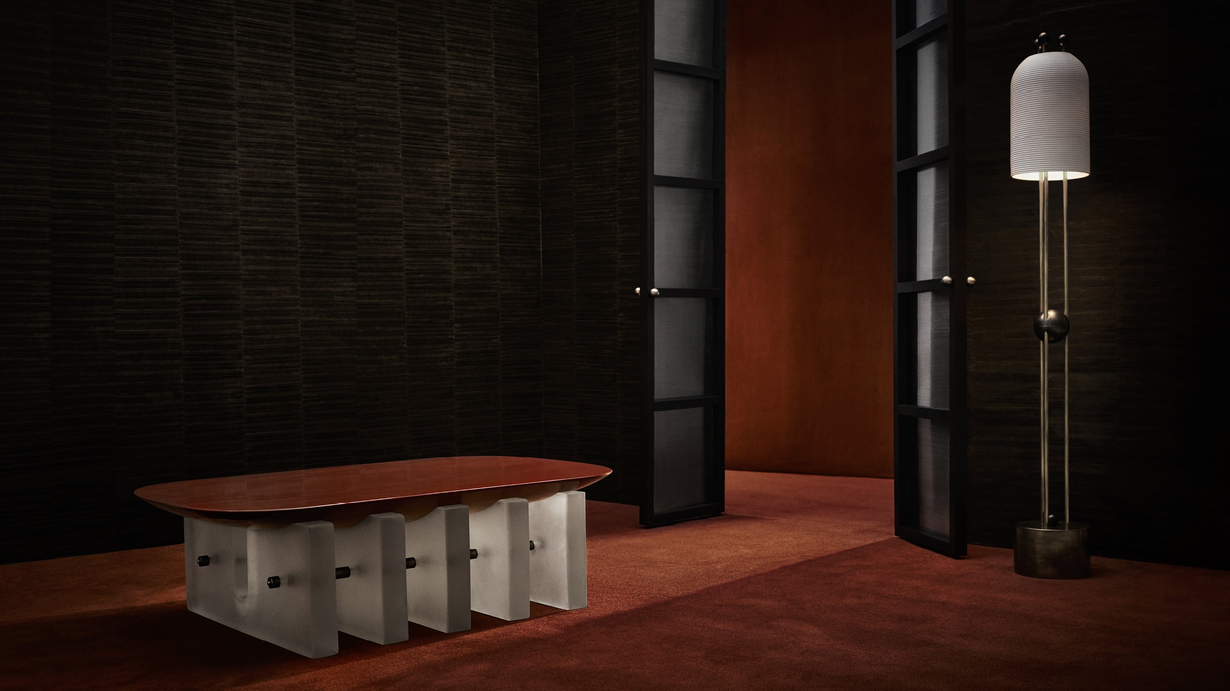 Minimalist interior with an oval wooden table on white slab bases and a tall cylindrical floor lamp by black paneled doors