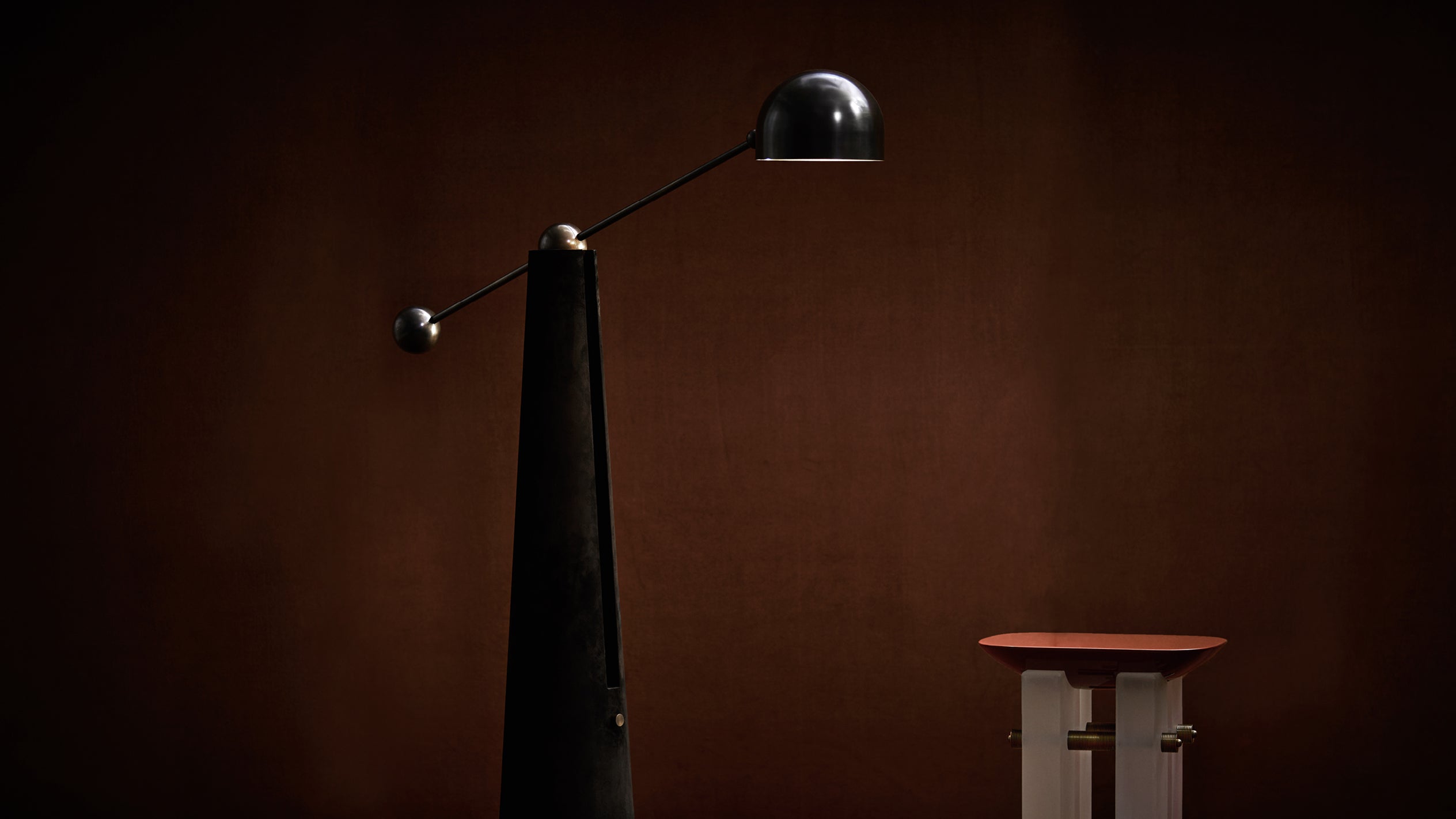 Black matte arched floor lamp with spherical counterweight next to small terracotta topped side table on brown background