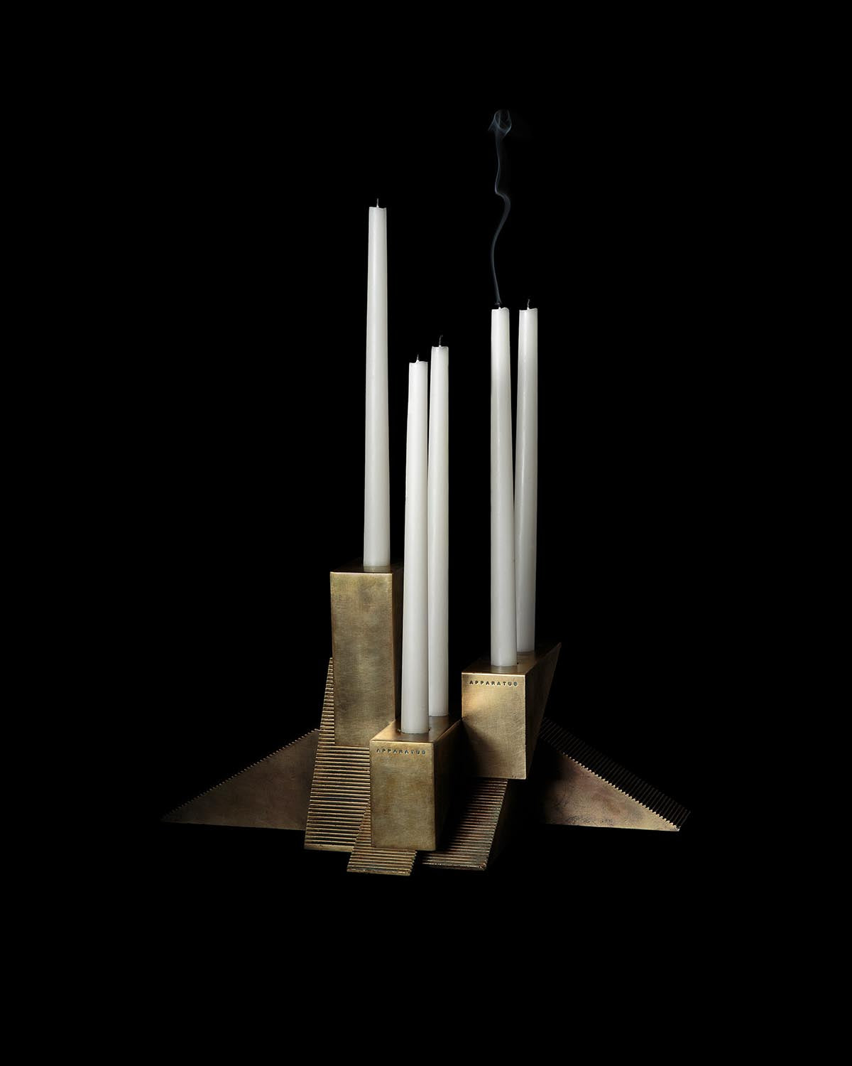 Geometric brass candle holder with multiple tall white taper candles against a black background
