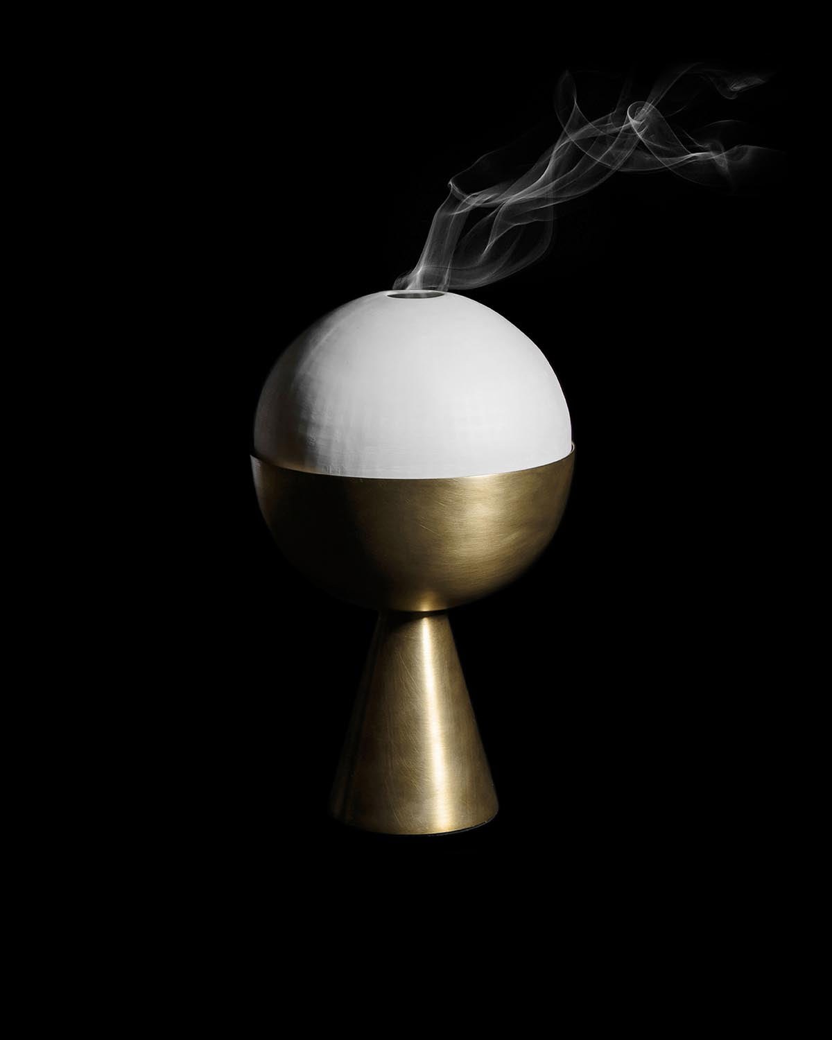 Spherical incense burner with matte white top and brushed brass base emitting thin wisps of smoke on a black background