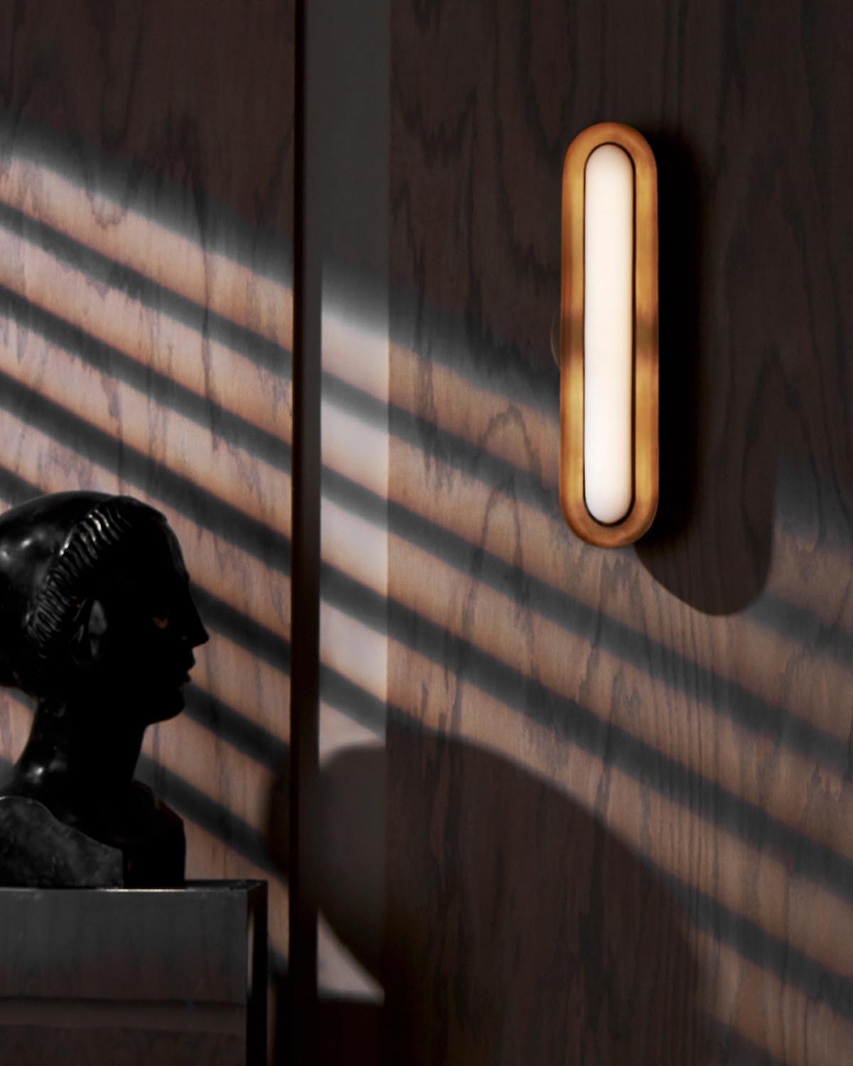 Vertical oval wall light with wooden frame casting shadows on textured wood panel wall above dark sculpture bust