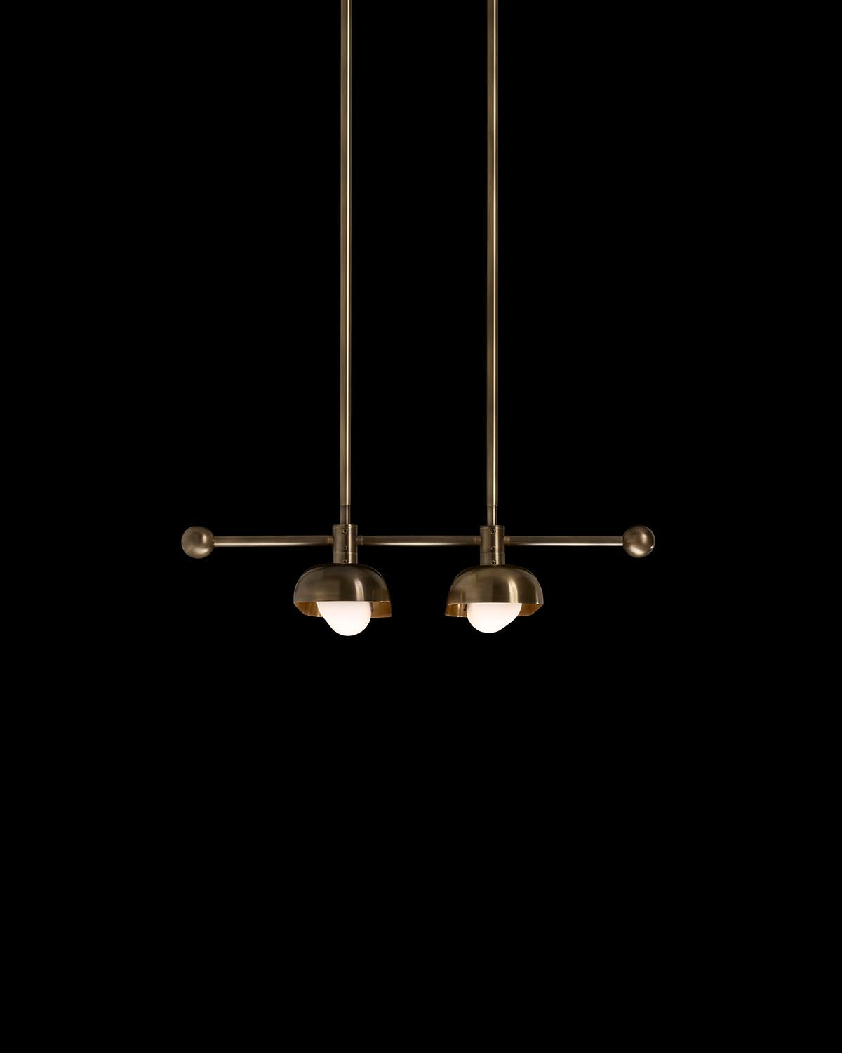 Minimalist dual pendant light fixture with round brass shades and exposed bulbs suspended against a black background