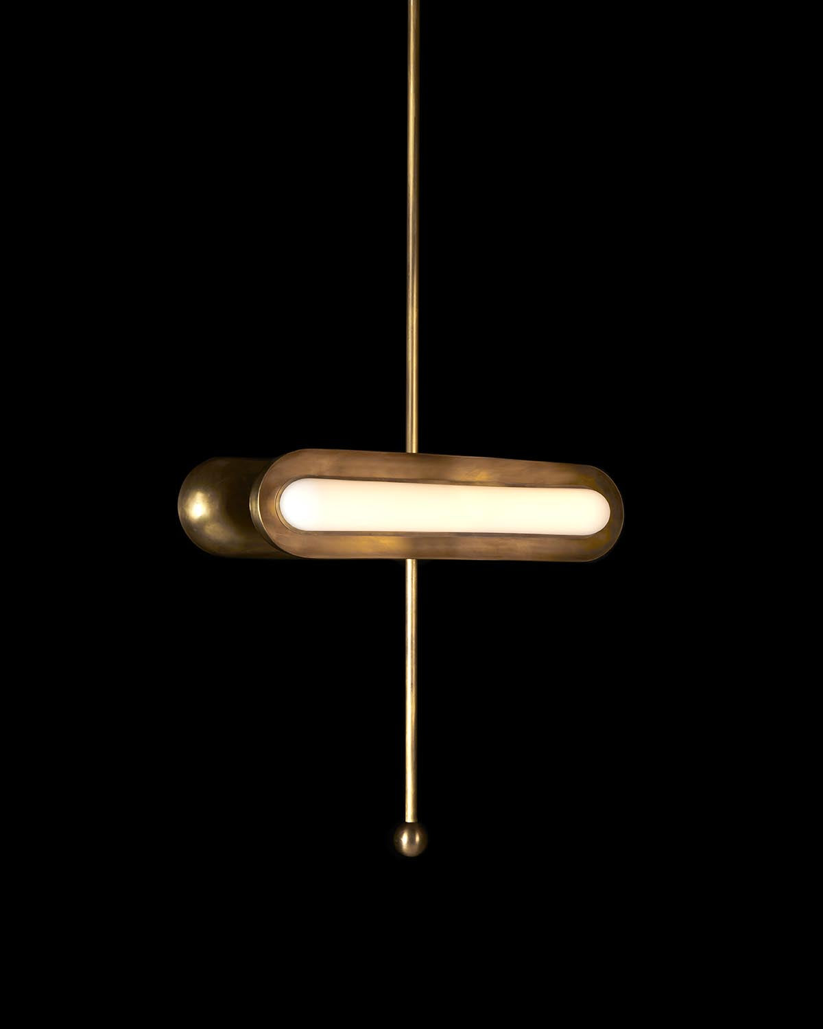 Horizontal brass pendant light with elongated frosted glass shade suspended on a thin vertical rod against black background